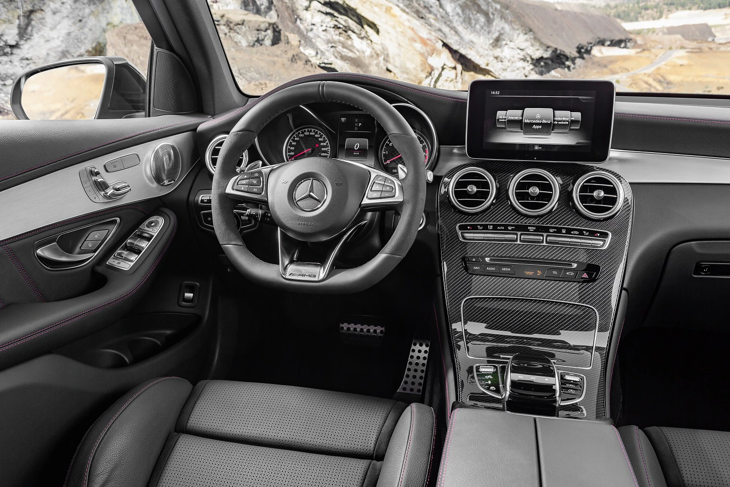 Innenraum eines modernen Mercedes-Autos mit Lederlenkrad, digitalem Display, Klimasteuerungen und schwarzen Ledersitzen. Draußen sind Felsen sichtbar.