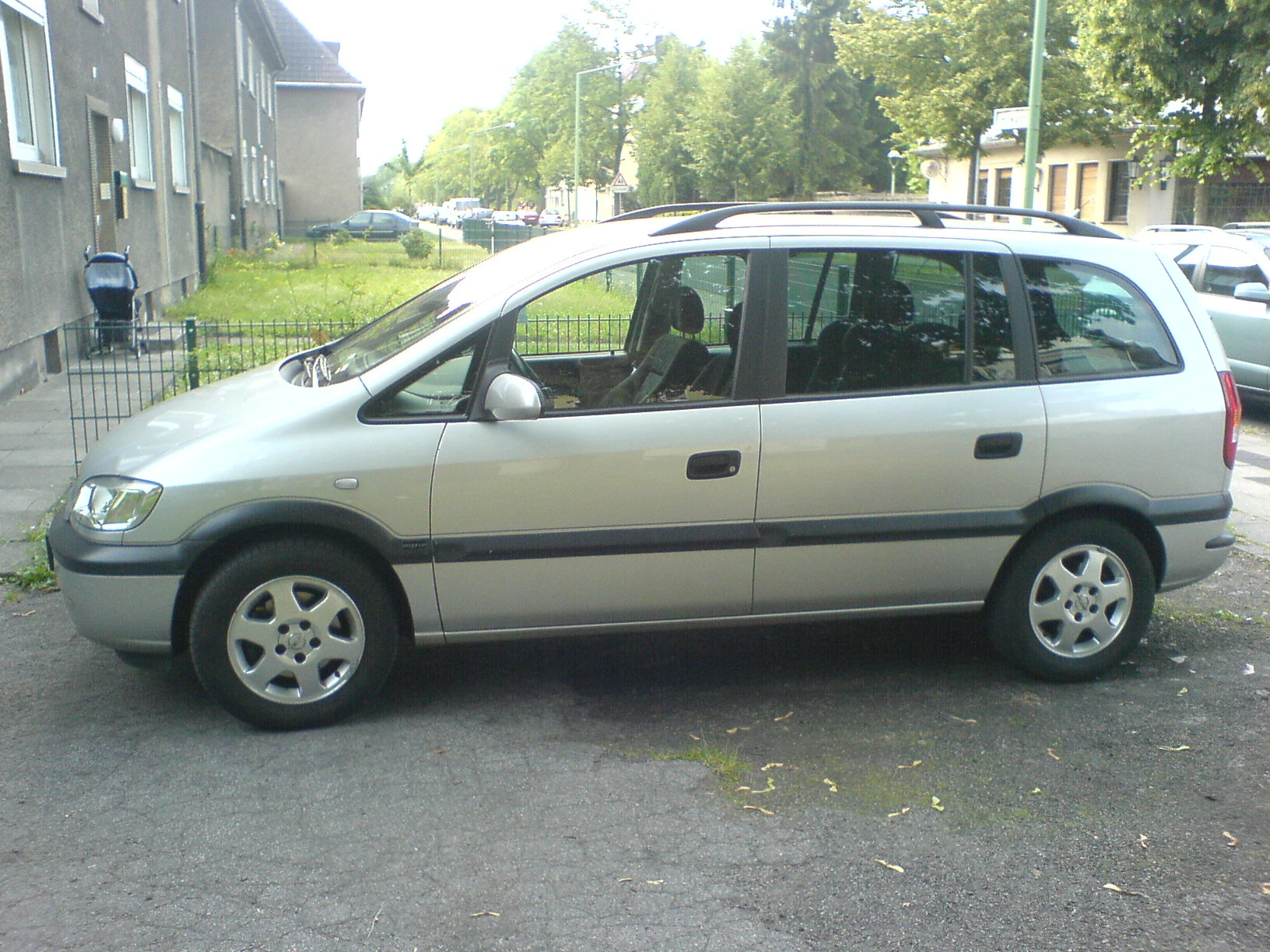 Silberner Minivan parkt auf einer Straße mit grüner Umgebung, links ein Kinderwagen und im Hintergrund Wohnhäuser.