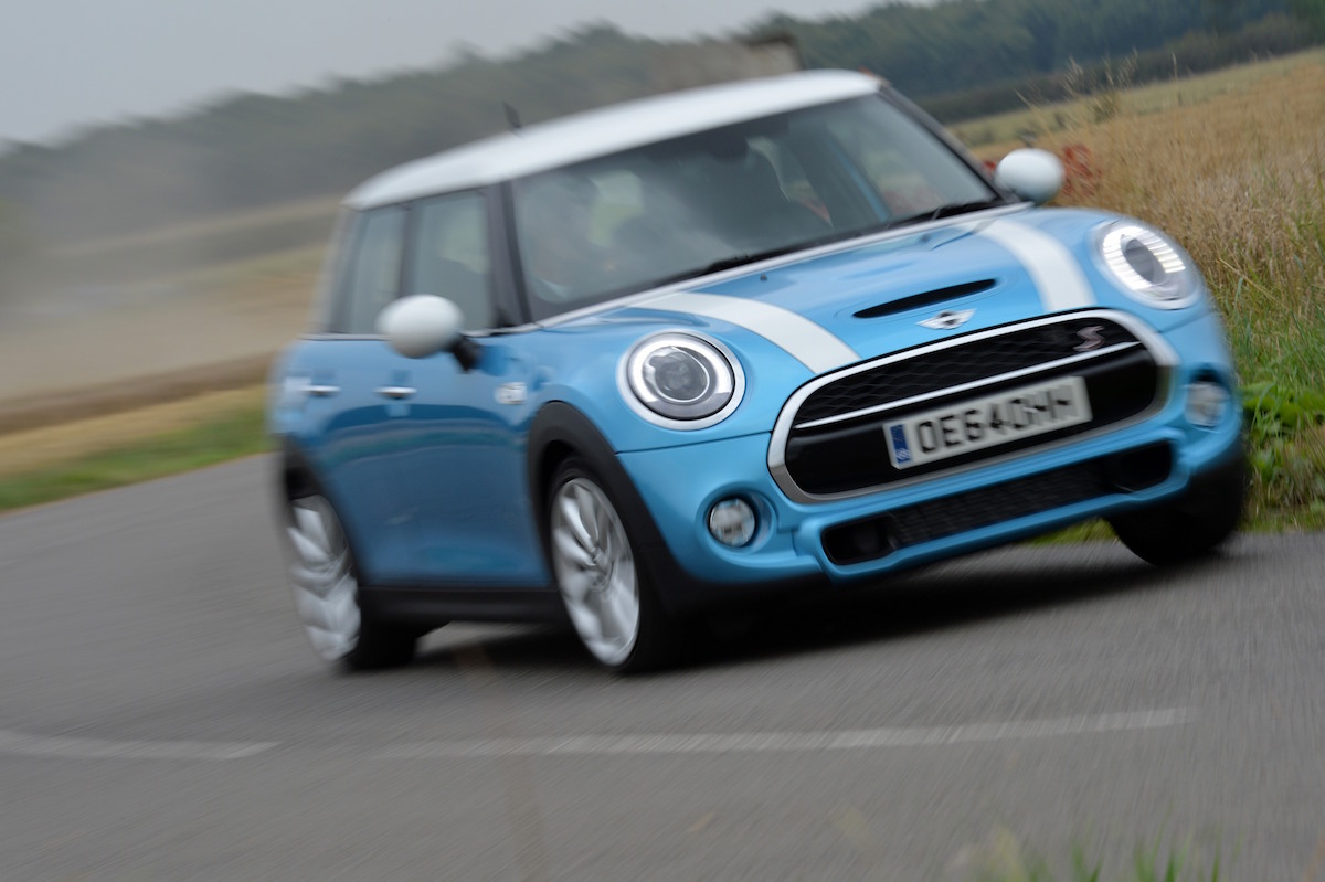 Blauer Mini Cooper fährt schnell um eine Kurve auf einer Landstraße, mit verschwommenem Hintergrund und ländlicher Umgebung.