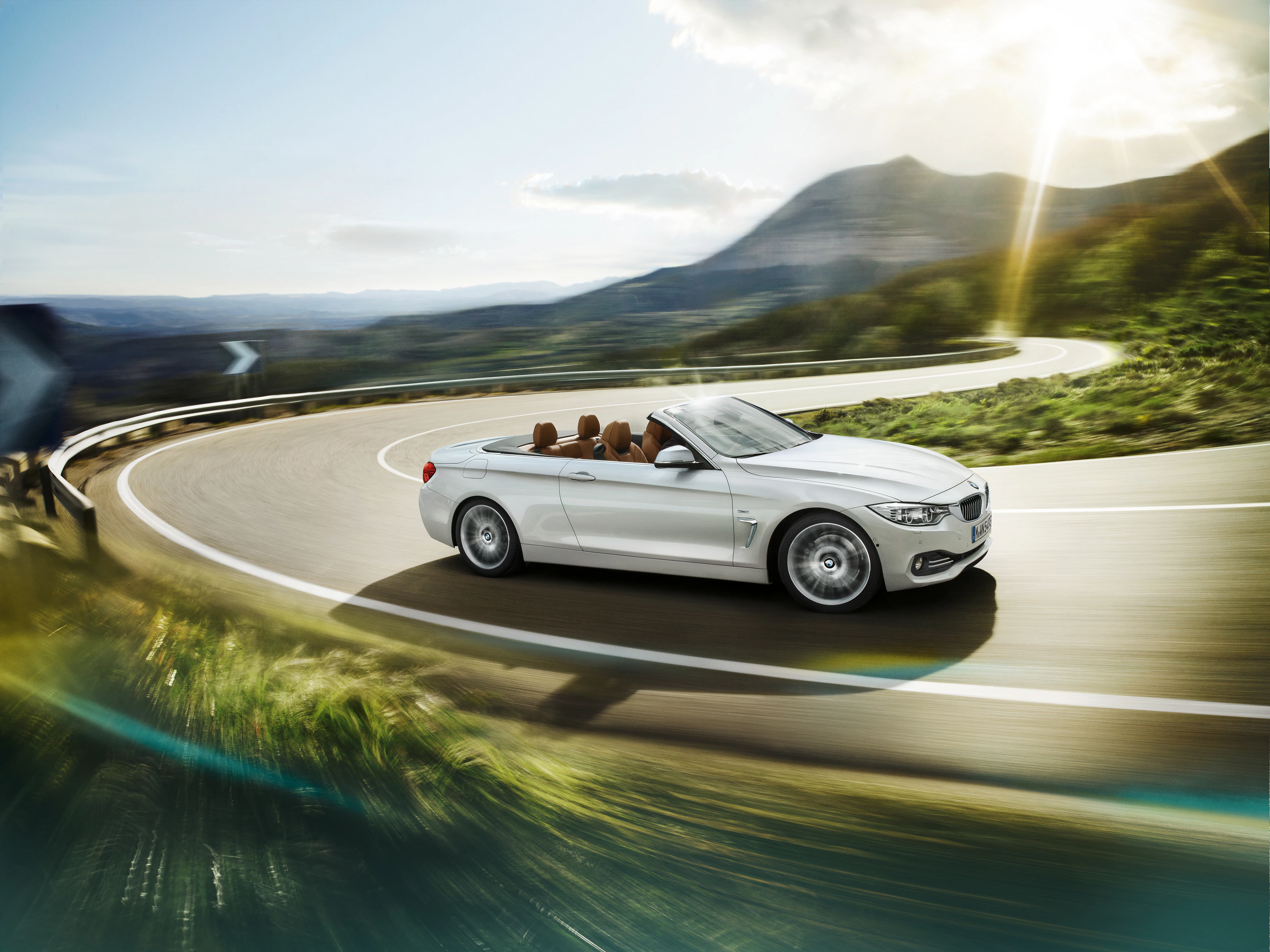Weißes Cabrio fährt dynamisch auf kurviger Landstraße bei Sonnenschein, umgeben von grüner Landschaft und Bergen im Hintergrund.
