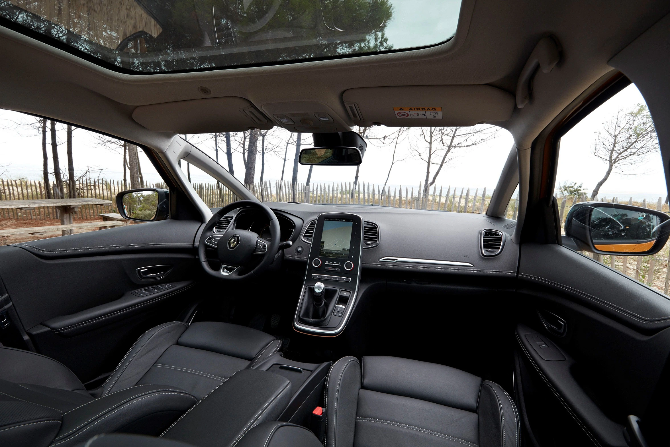 Innenansicht eines Autos mit Glasdach, Blick auf Lenkrad, Mittelkonsole und Ledersitze, im Hintergrund Bäume und Holzzaun.