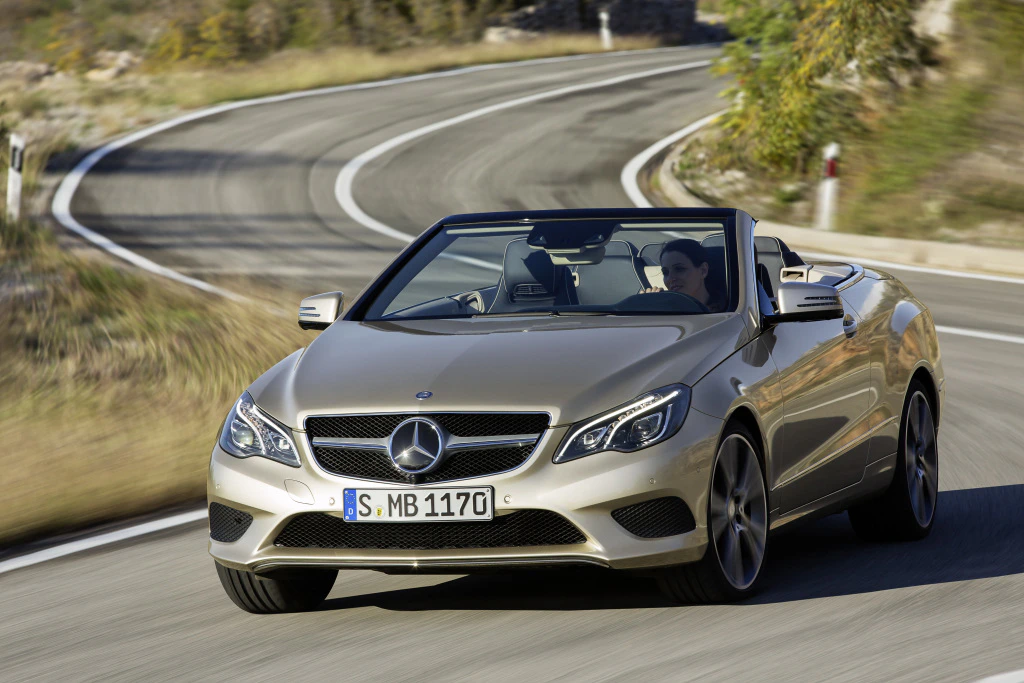 Beiges Mercedes-Benz Cabrio in Kurve auf Landstraße, Sonnenschein, Fahrer am Steuer.