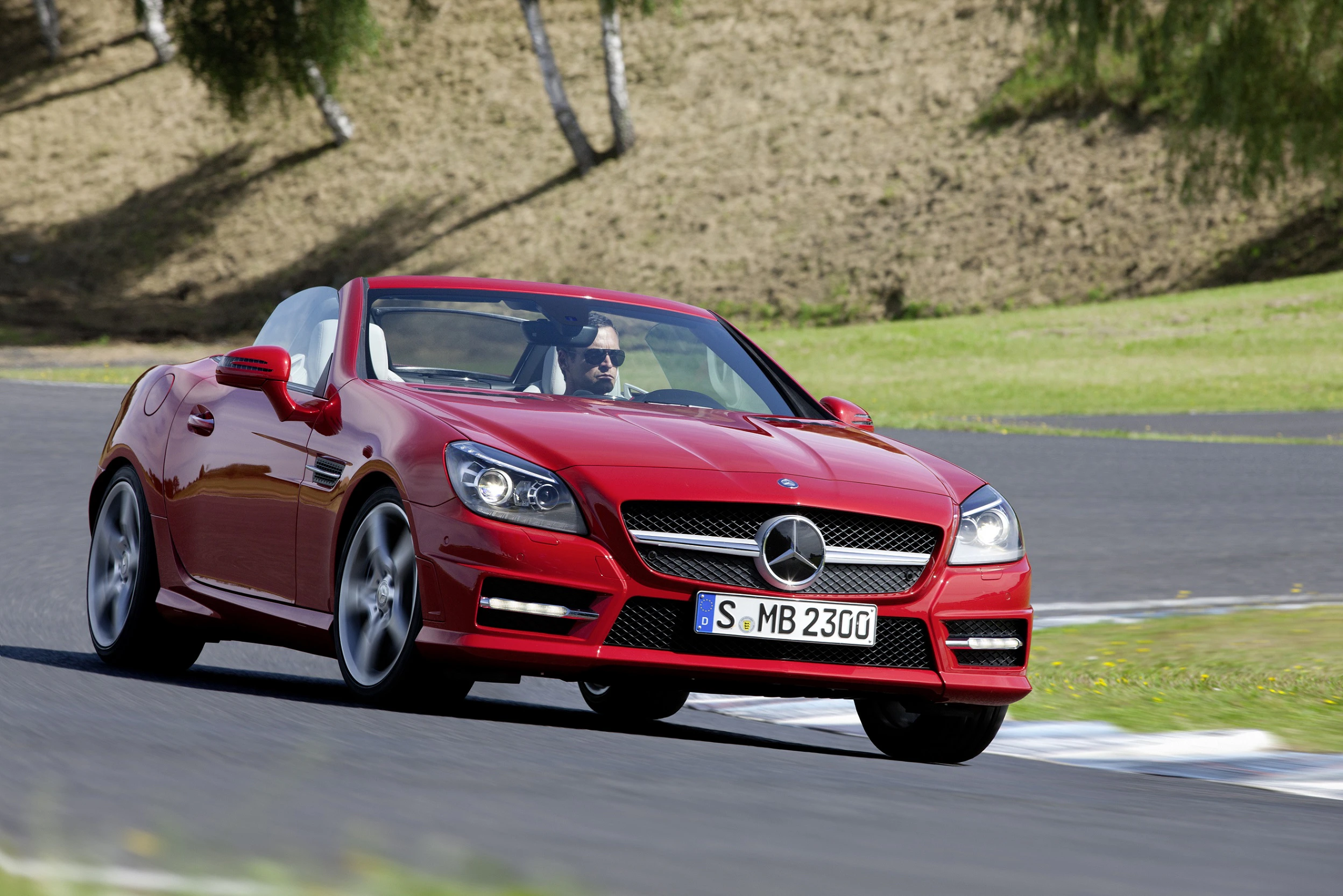 Roter Mercedes-Cabrio fährt auf einer kurvenreichen Straße; klare Himmel, Bäume im Hintergrund, Fahrer mit Sonnenbrille am Steuer.