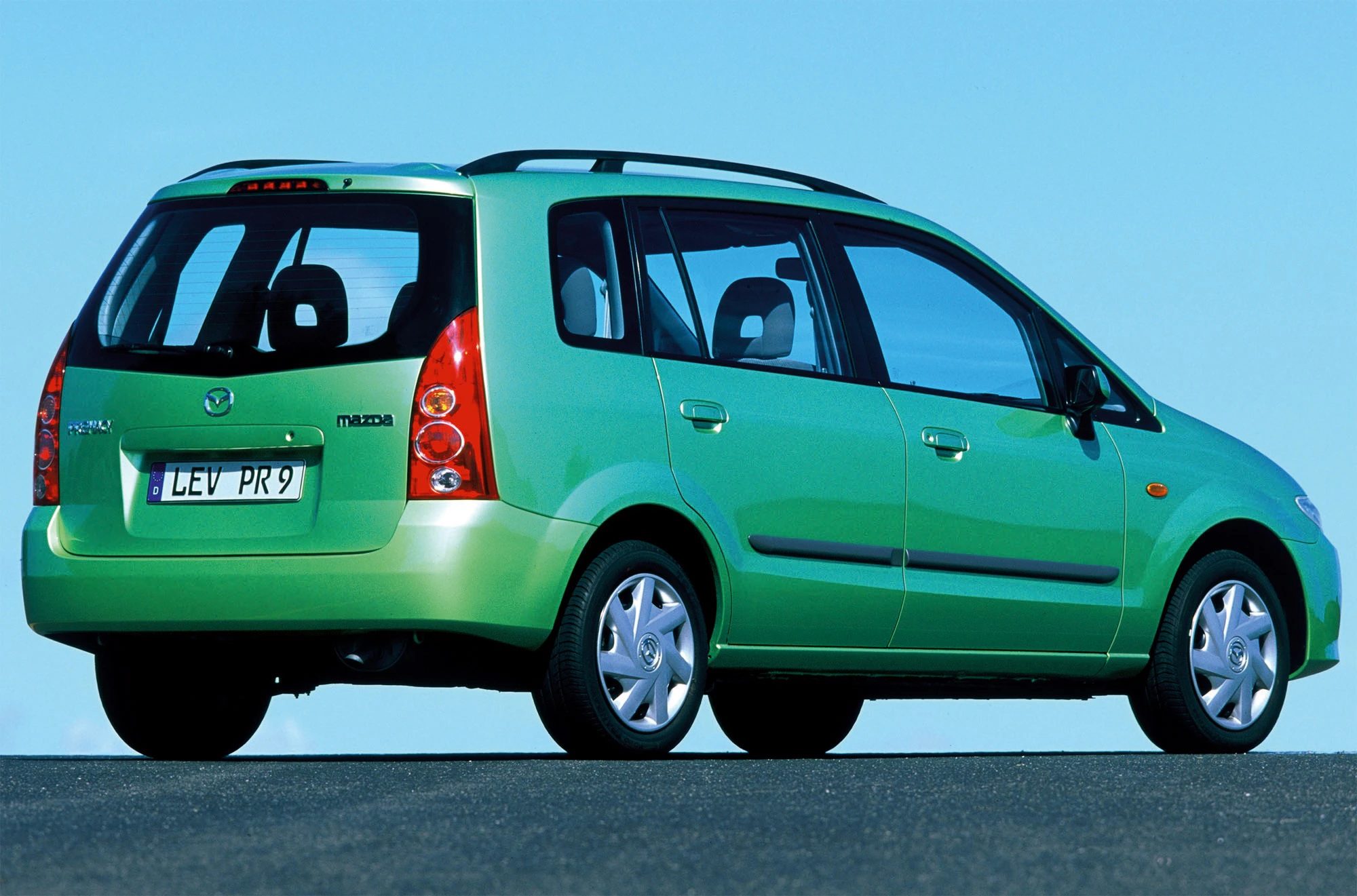 Grüner Mazda Premacy Minivan auf einer Straße mit blauem Himmel im Hintergrund, von hinten seitlich fotografiert.