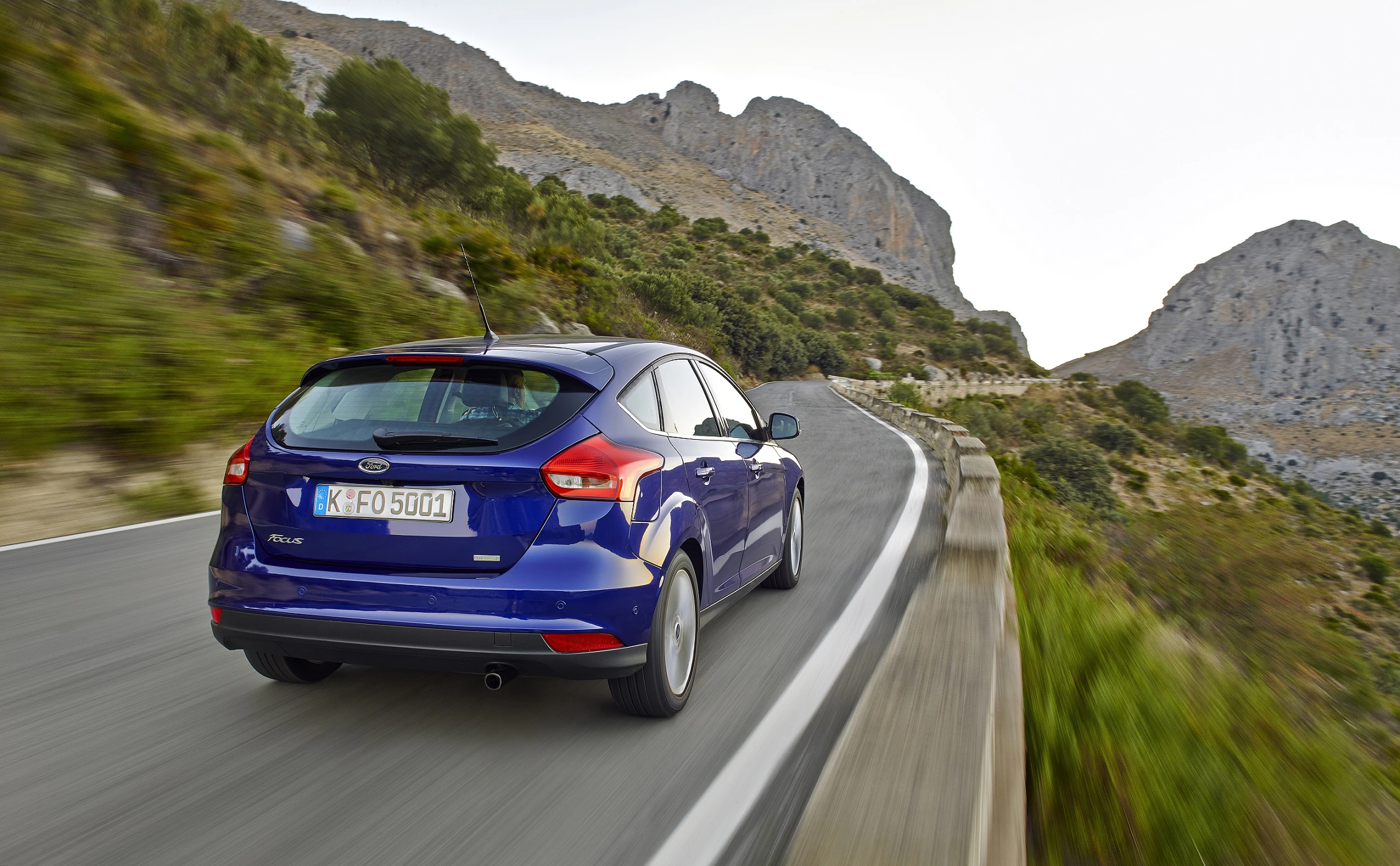 Blauer Ford fährt auf kurvenreicher Bergstraße, umgeben von grüner Vegetation und felsigen Hügeln unter klarem Himmel.
