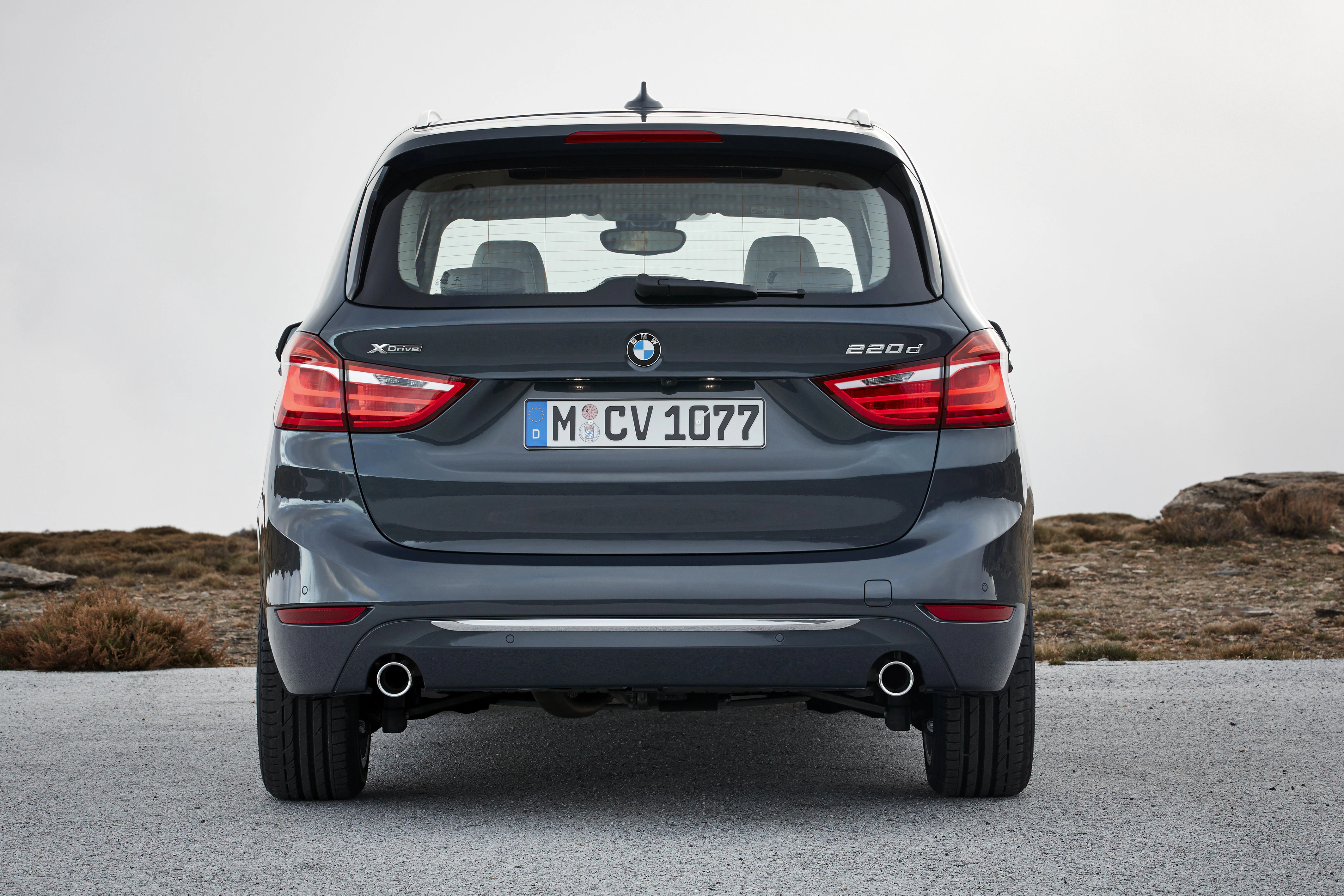 Rückansicht eines grauen BMW 220d auf einer Straße, Nummernschild "M CV 1077", in einer natürlichen Umgebung.