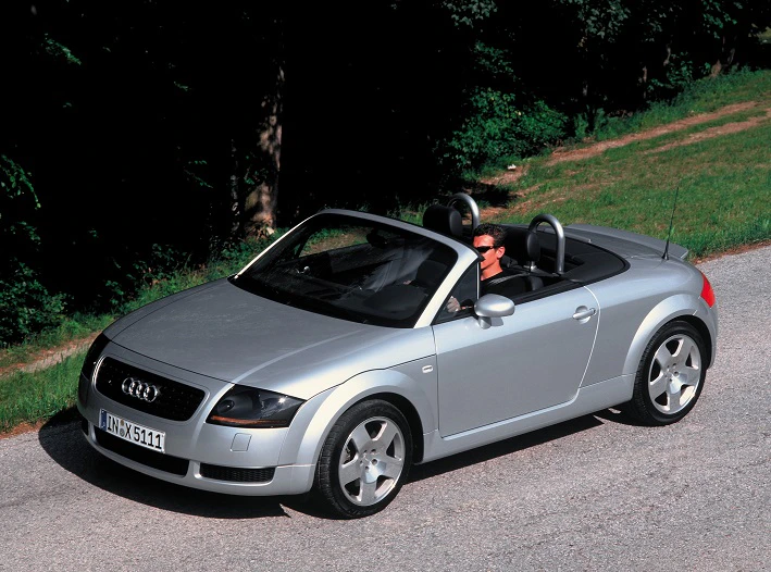 Silberner Audi TT Cabrio fährt auf ländlicher Straße, Fahrer trägt Sonnenbrille. Hintergrund aus Bäumen und Gras.