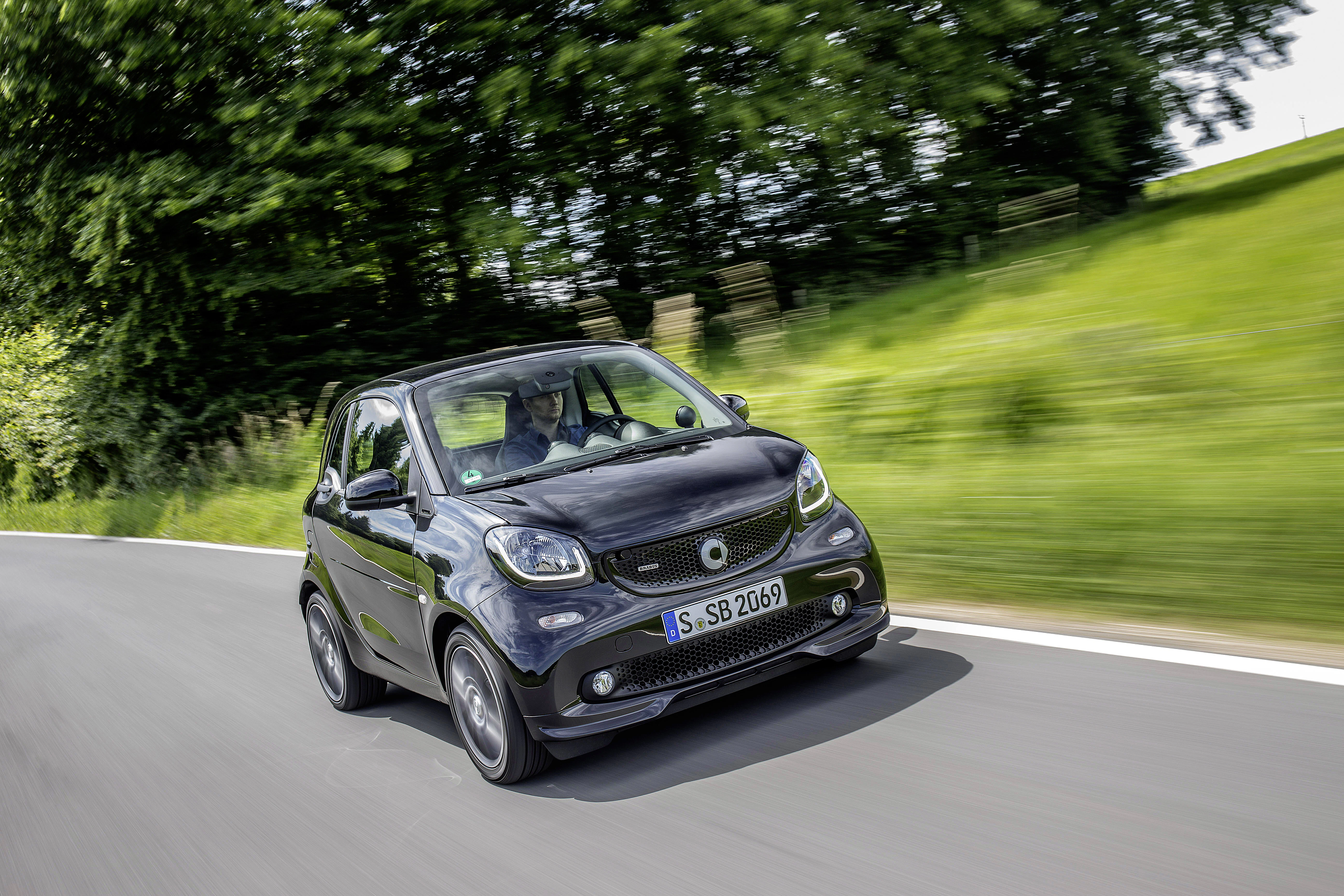 Kleines, schwarzes Auto fährt auf einer Landstraße, umgeben von grüner Landschaft und Bäumen. Fahrer im Fahrzeug.