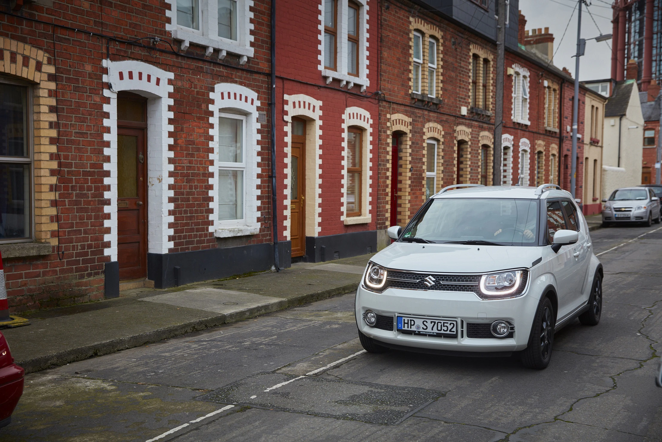 Weißer Suzuki Ignis fährt auf schmaler Straße mit viktorianischen Backsteinhäusern im Hintergrund.