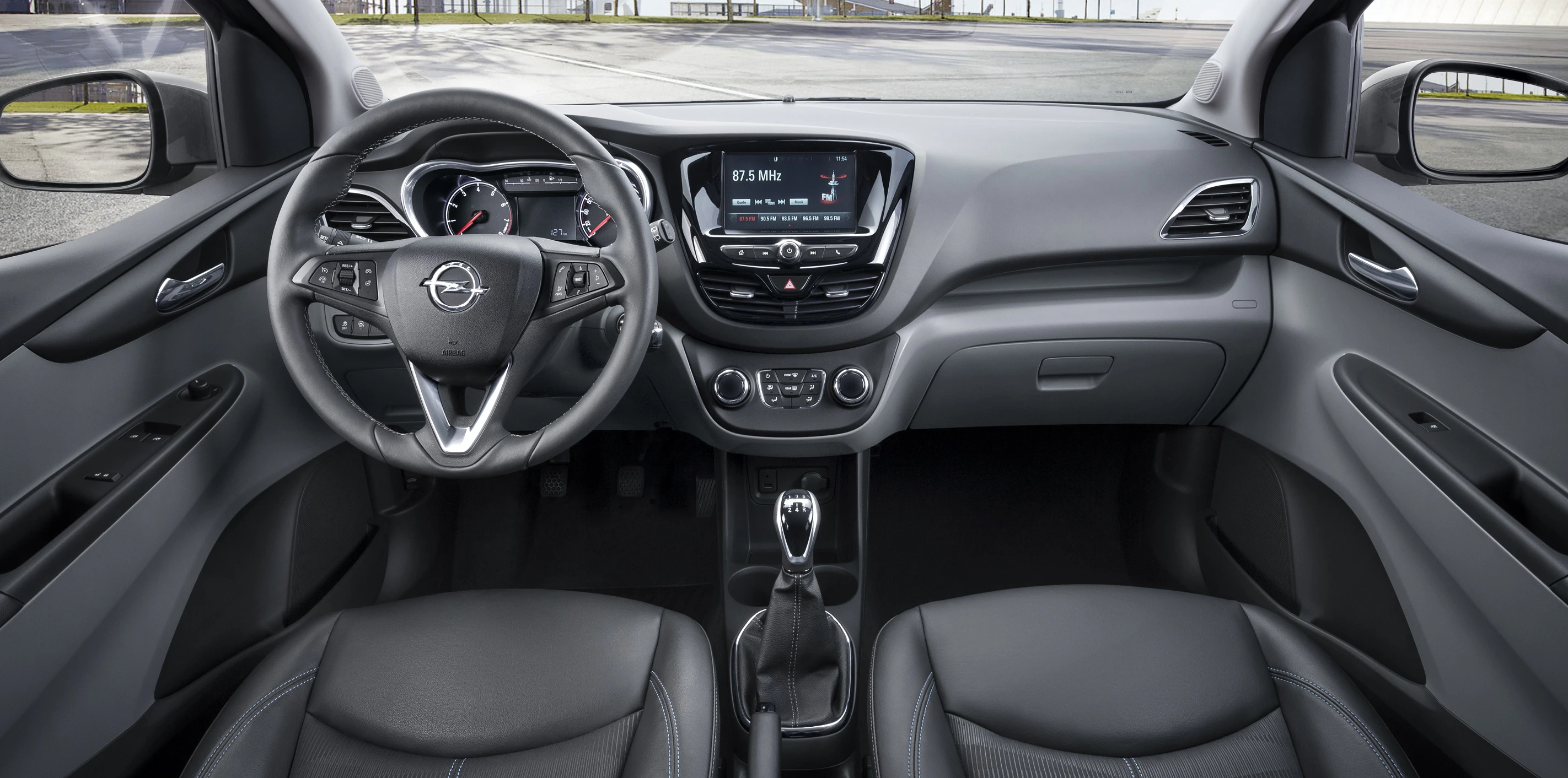 Innenraumansicht eines modernen Autos mit Lenkung, Mittelkonsole, Touchscreen-Display und Schalthebel, umgeben von Ledersitzen.