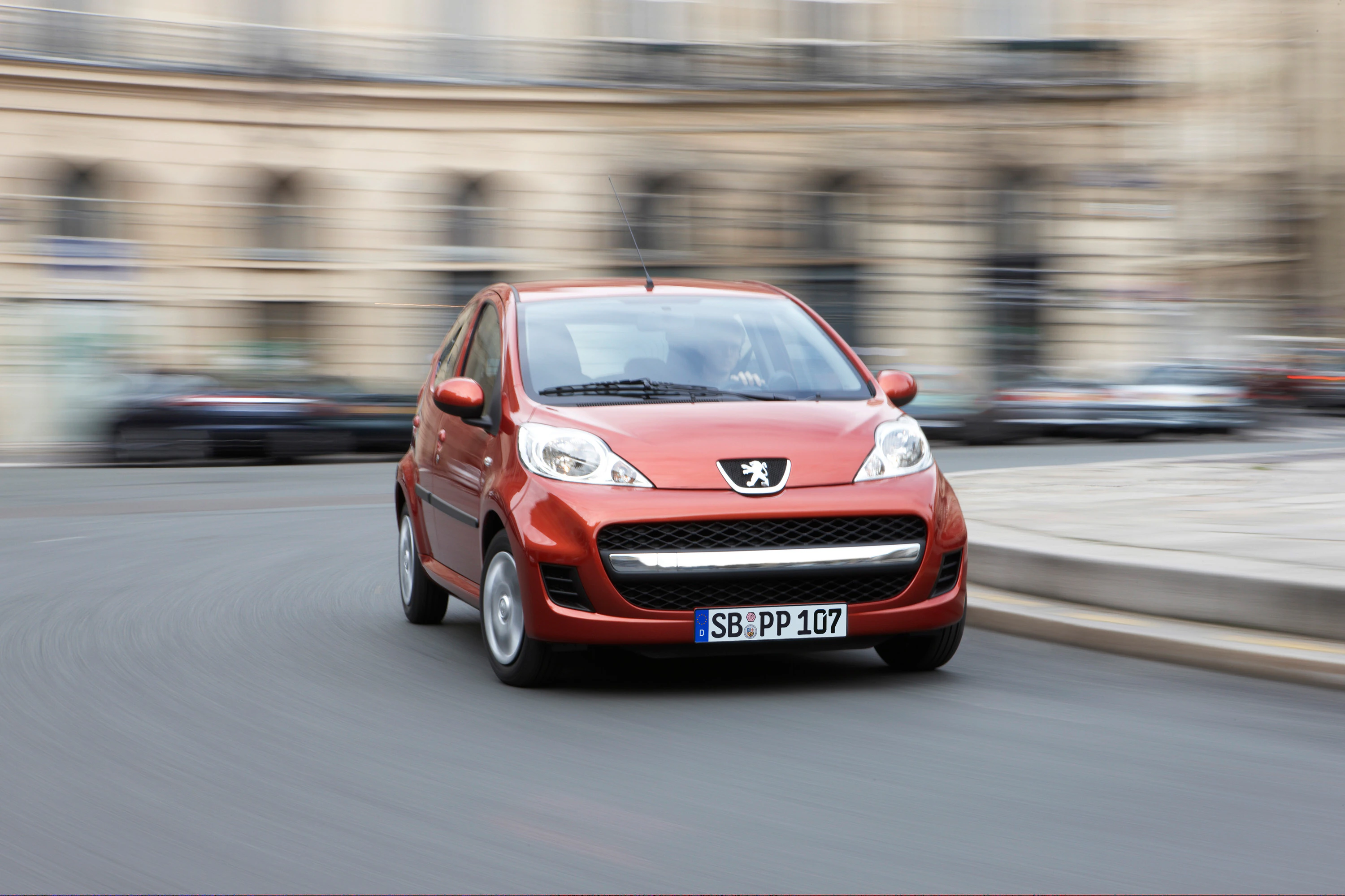 Roter Peugeot 107 fährt auf einer städtischen Straße. Gebäude im Hintergrund sind unscharf, was die Bewegung des Autos betont.