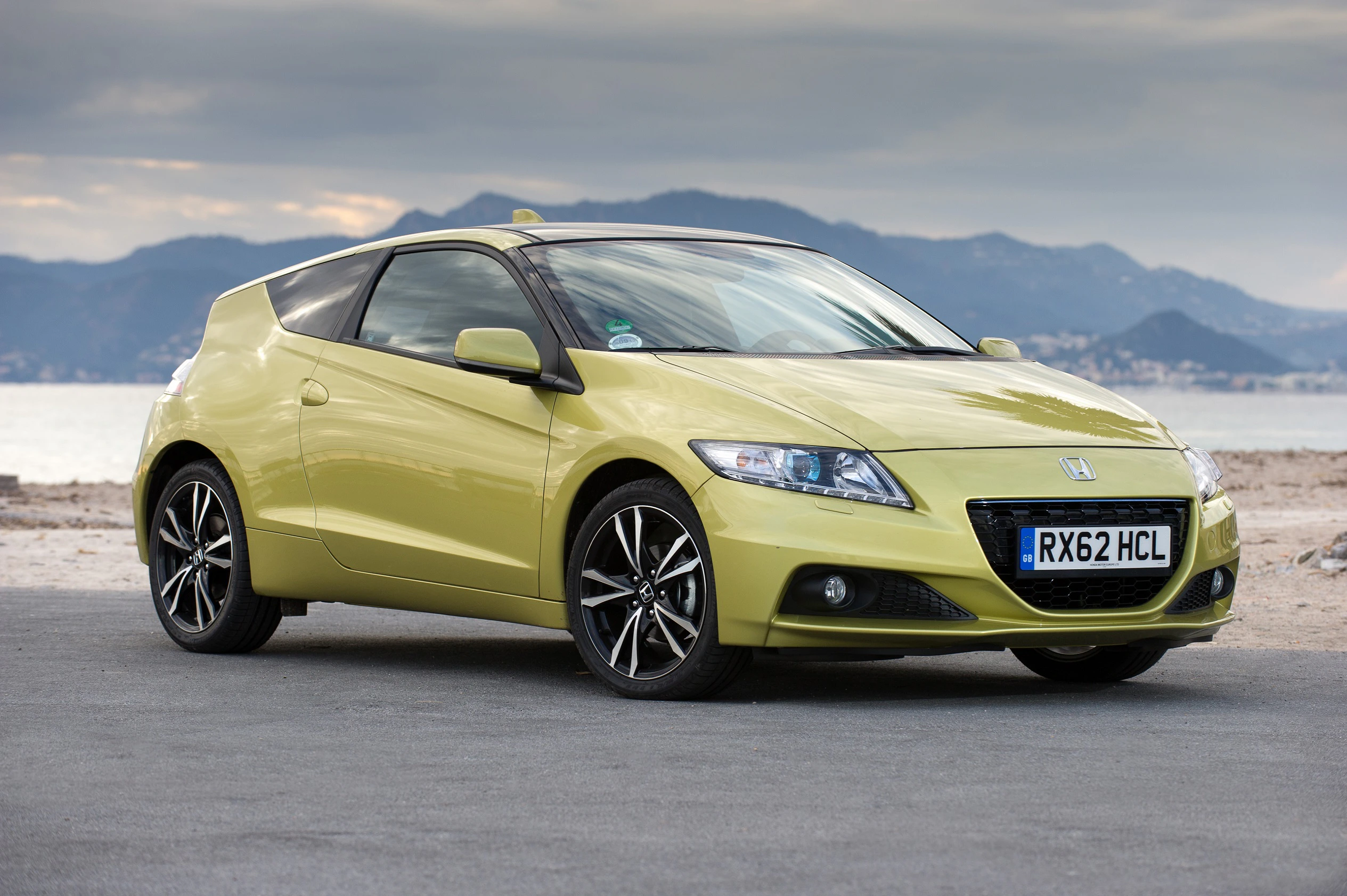 Hellgrüner Honda CR-Z auf einer Straße, im Hintergrund ein Meer und Berge unter bewölktem Himmel.