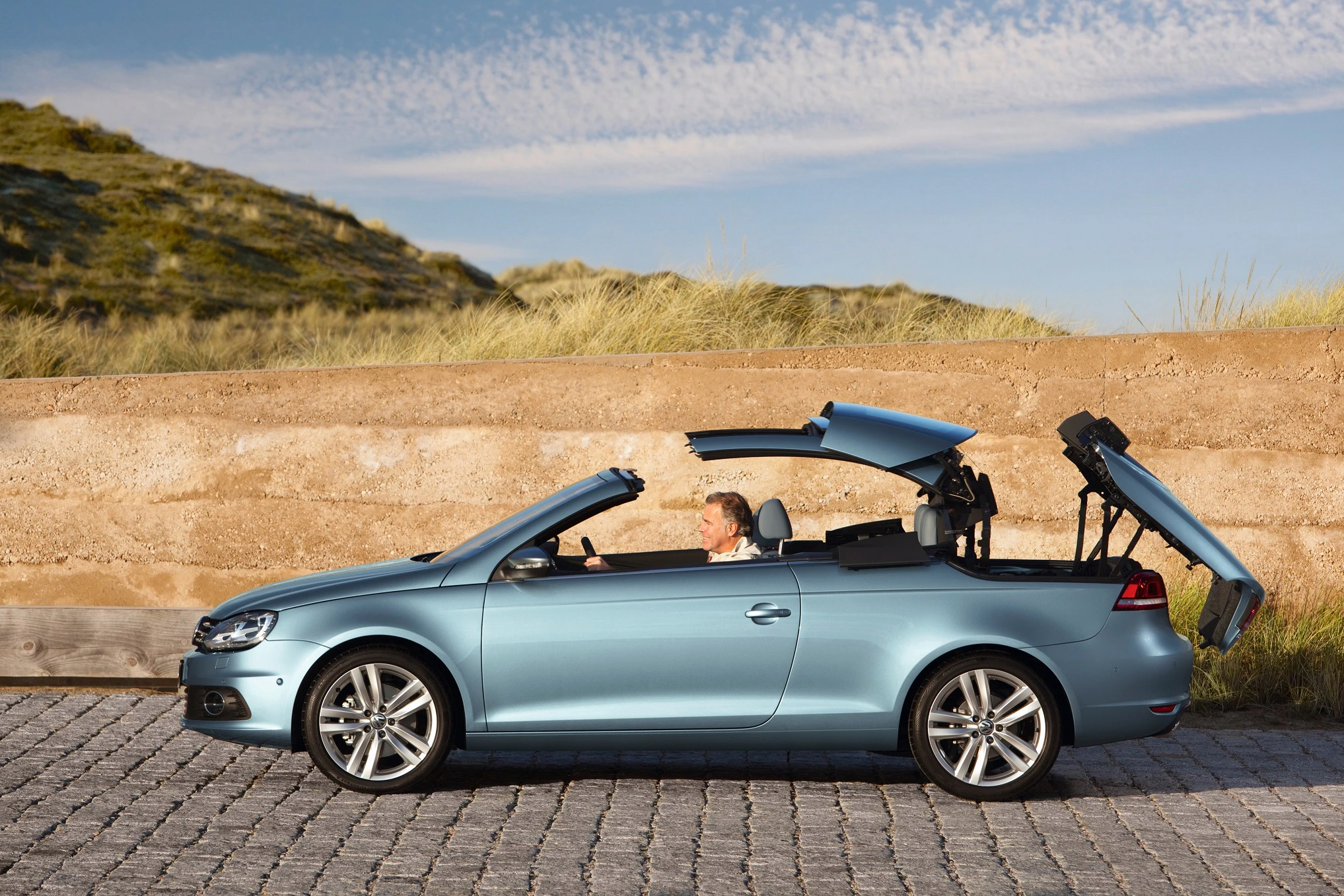 Ein blauer Cabrio mit geöffnetem Dach, geparkt vor einer Steinmauer und Dünen. Fahrer sitzt am Steuer auf einer gepflasterten Straße.