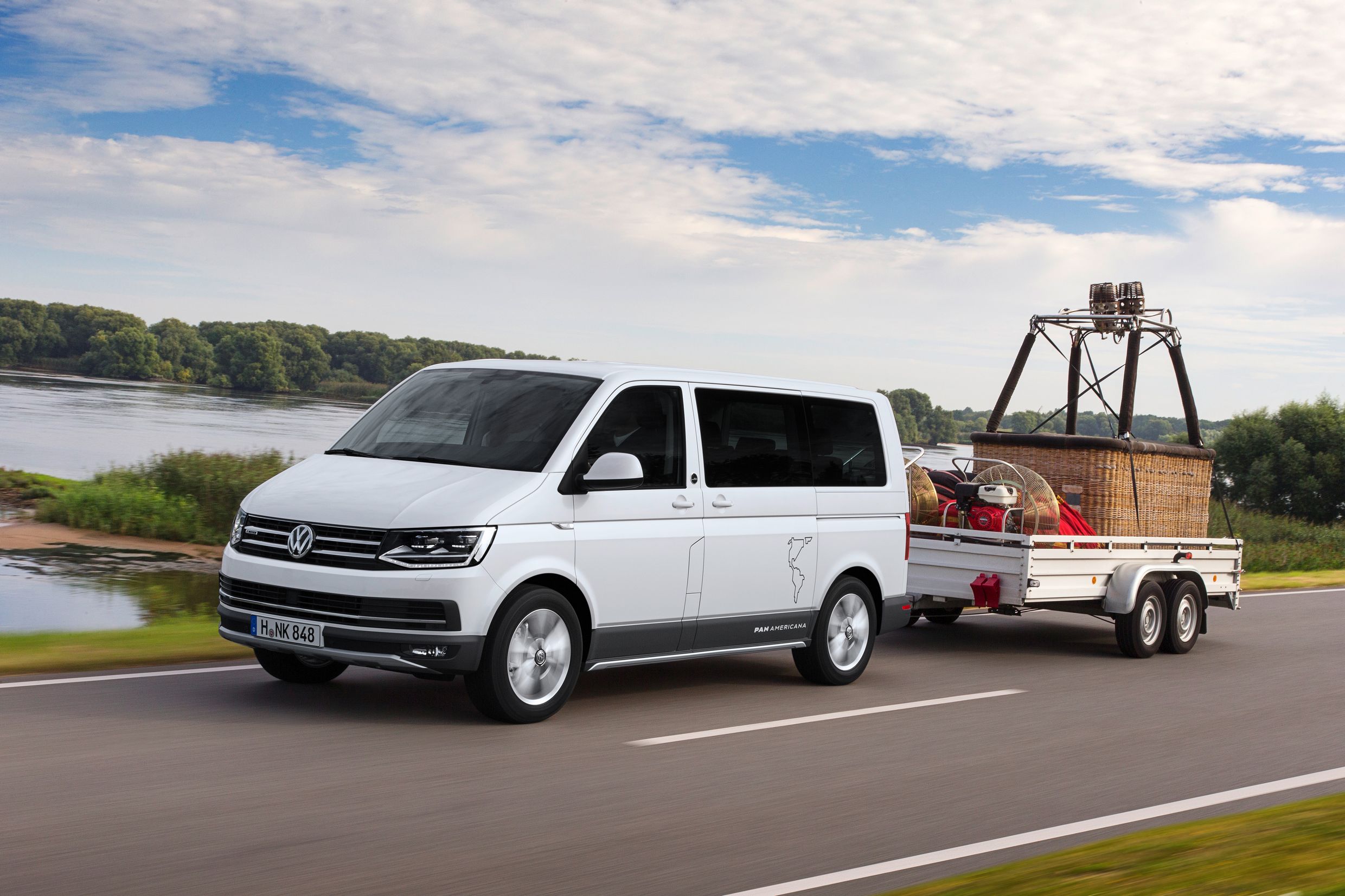 Weißer VW-Bus zieht Anhänger mit Heißluftballonausrüstung auf einer Landstraße vor einem Fluss und grüner Landschaft.