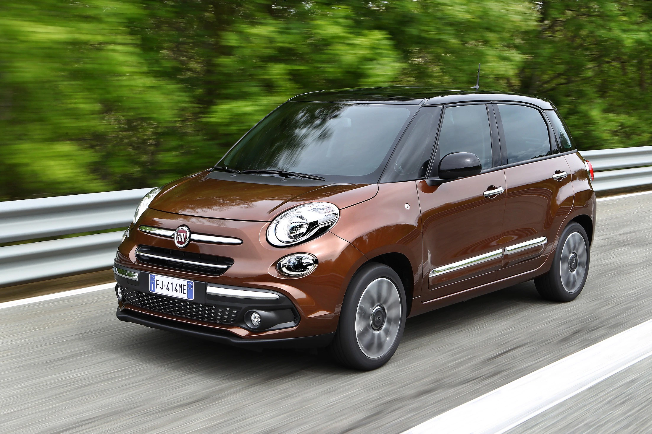 Brauner Fiat 500L fährt auf einer Landstraße mit Bäumen im Hintergrund. Dynamische Bewegung und schlankes Design hervorgehoben.
