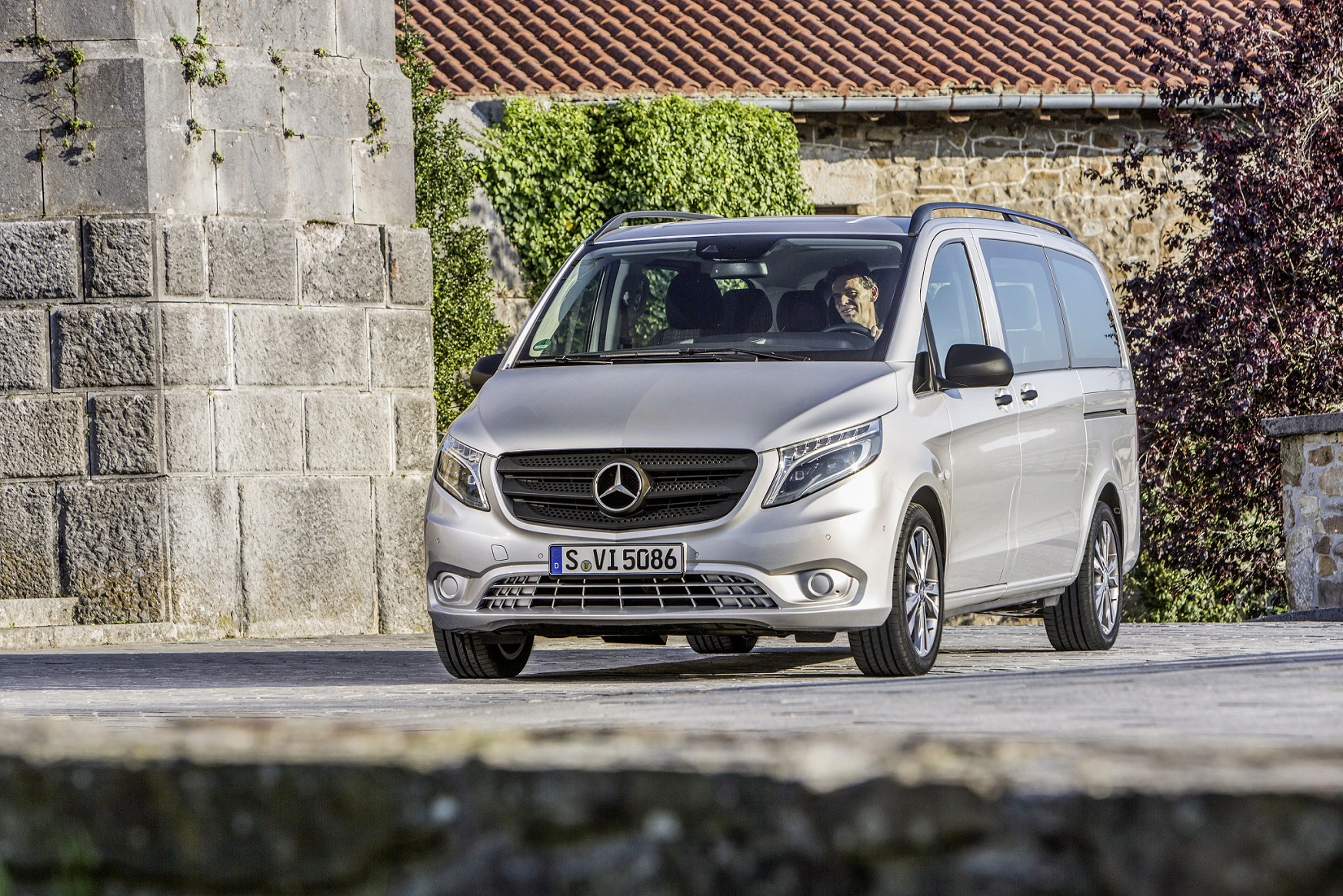 Silberner Mercedes-Benz Vito parkt vor einem historischen Gebäude, rote Ziegeldächer und alte Steinwände im Hintergrund.