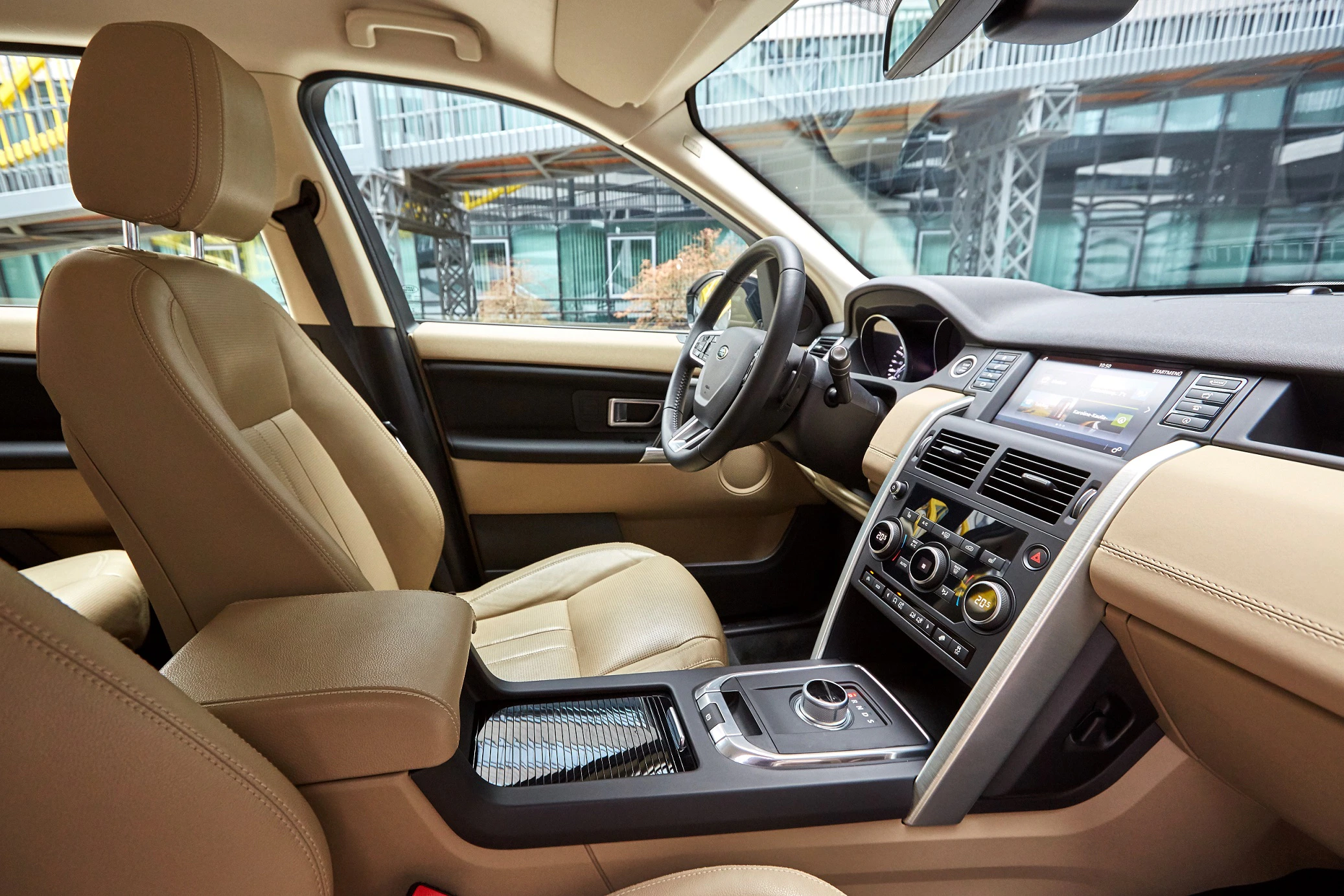 Innenraum eines Autos mit beigen Ledersitzen, modernem Armaturenbrett, Touchscreen, Lenkrad und verschiedenen Bedienelementen.