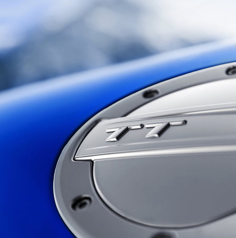 Nahaufnahme eines Autotankdeckels mit "TT"-Logo auf glänzendem Metall, umgeben von blauer Karosserie. Im Hintergrund verschwommene Berge.