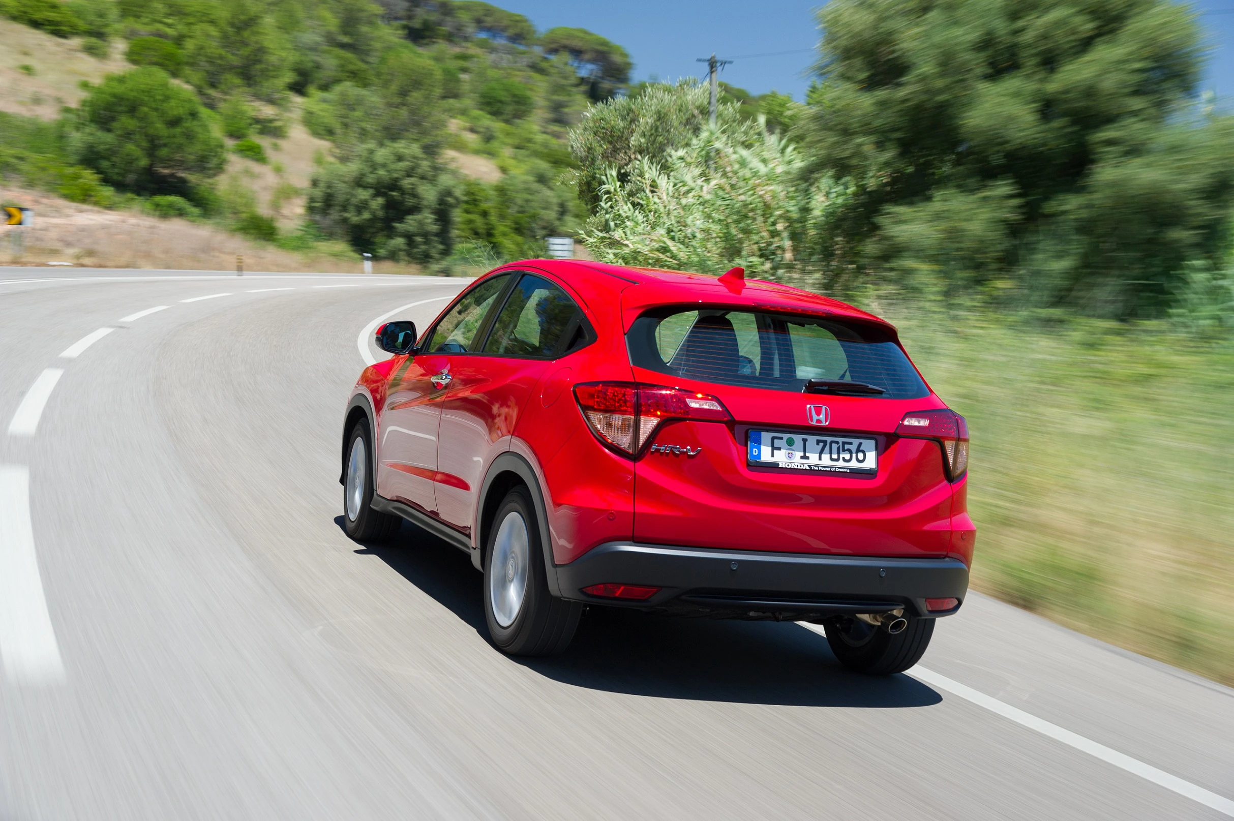 Roter Honda HR-V fährt auf einer kurvenreichen Landstraße bei sonnigem Wetter, umgeben von grüner Vegetation.