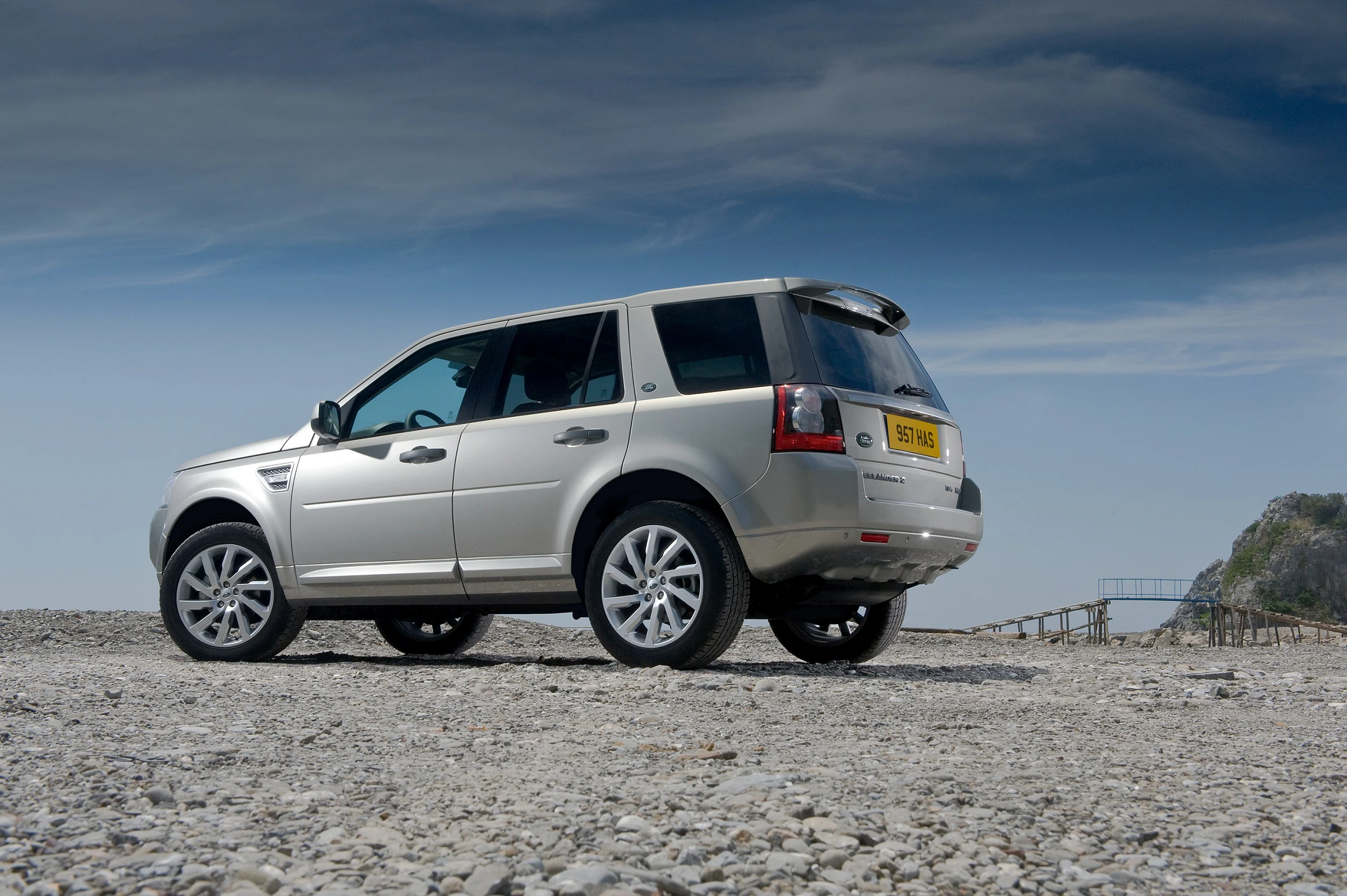 Silberner SUV auf Kiesstrand, blauer Himmel im Hintergrund. Fahrzeug parkt an abgelegenem, landschaftlich reizvollem Ort.