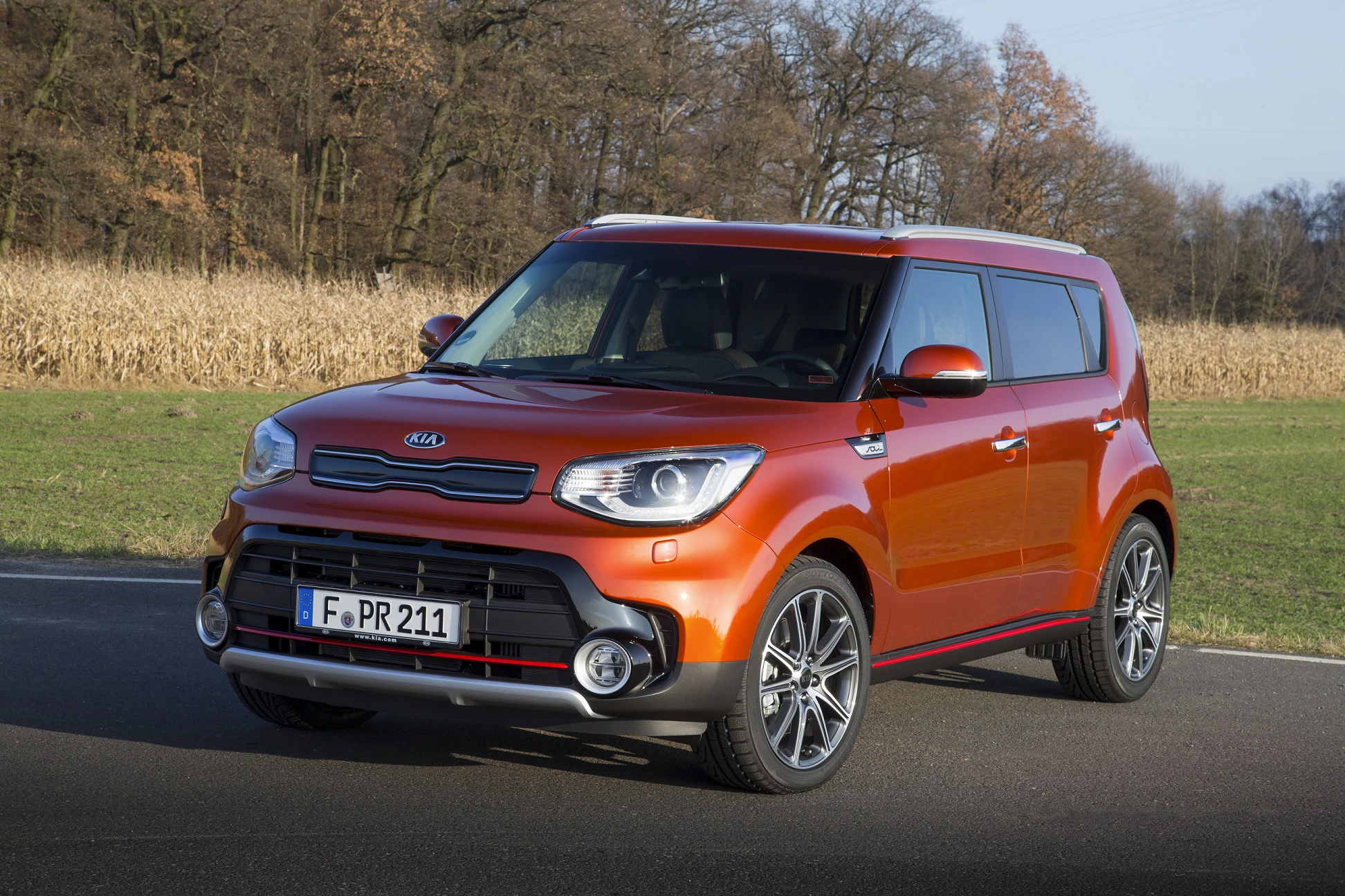 Ein orangefarbener Kia Soul steht auf einer Straße, umgeben von Bäumen und einem Maisfeld im Hintergrund.