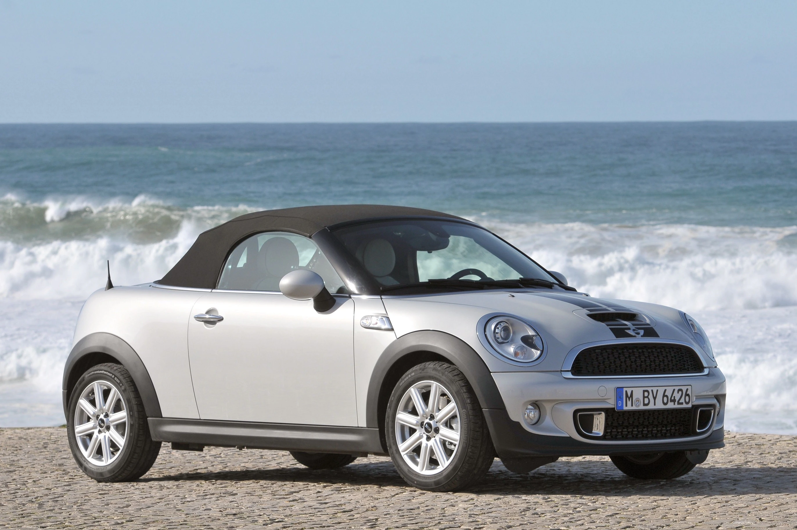Silberner Mini Cooper Cabrio parkt am Strand mit Meereswellen im Hintergrund. Dach geschlossen, sonniger Tag.