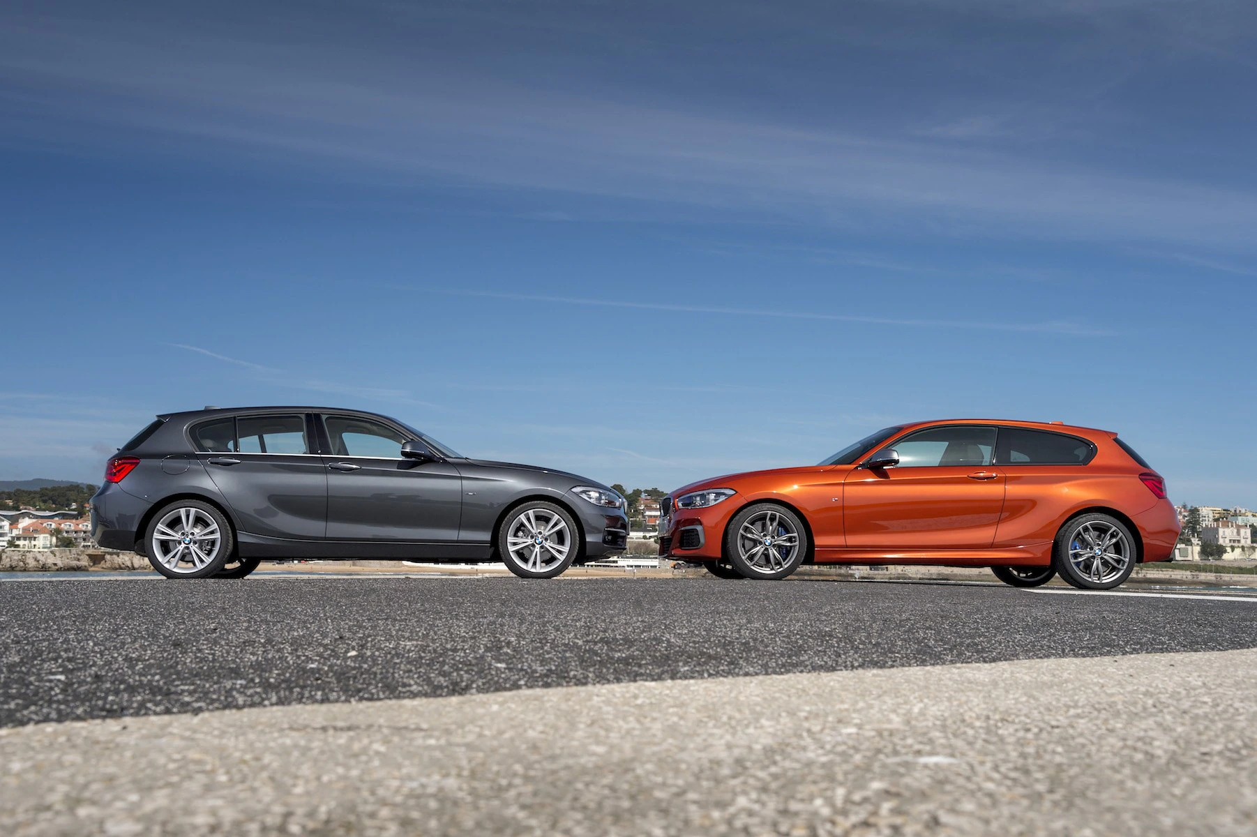 Zwei Autos stehen sich auf einer Straße gegenüber, eines ist grau, das andere orange. Der Himmel ist blau und klar im Hintergrund.