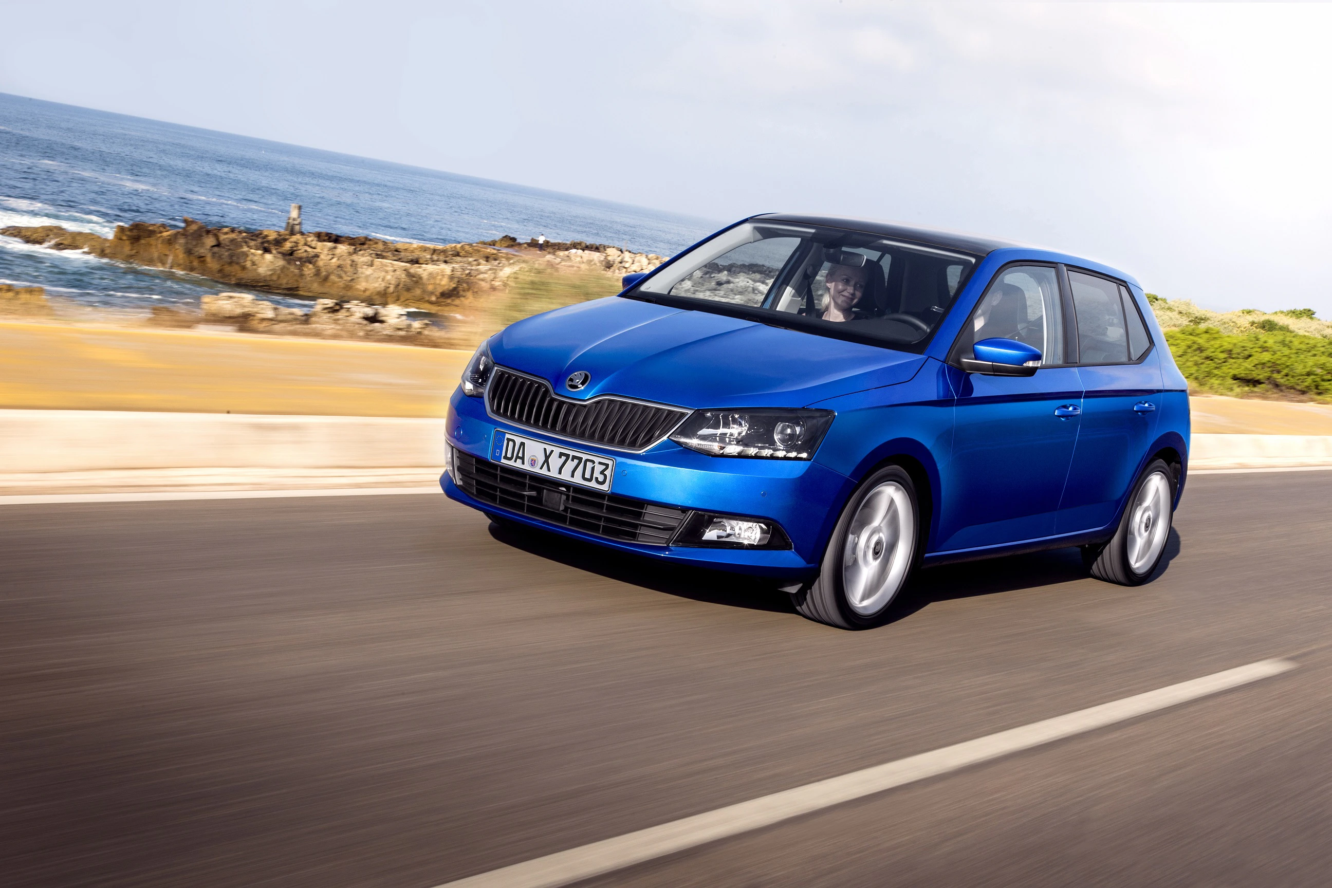 Blauer Skoda Fabia fährt auf Küstenstraße mit Meer im Hintergrund bei sonnigem Wetter. Fahrer am Steuer.