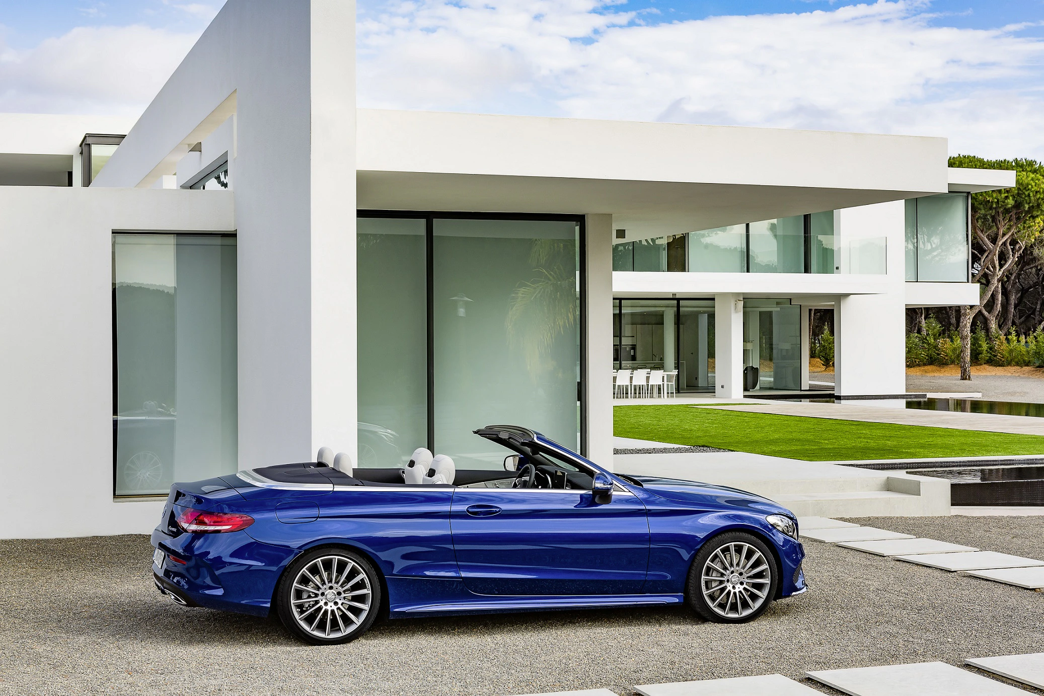 Blaues Cabrio vor modernem, weißem Haus mit großen Glasfenstern und gepflegtem Rasen, bei sonnigem Wetter geparkt.