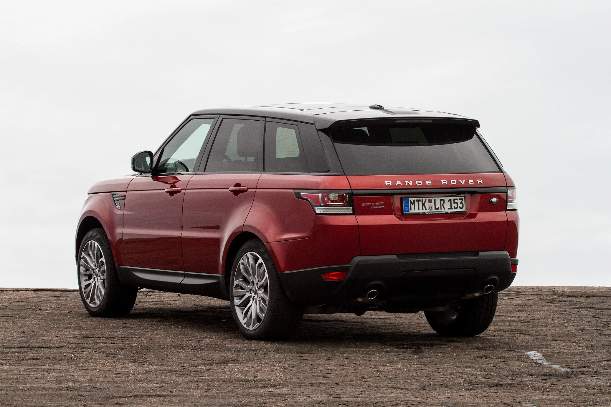Roter Range Rover SUV, geparkt auf grauem Asphalt vor bewölktem Himmel, von schräg hinten links fotografiert.