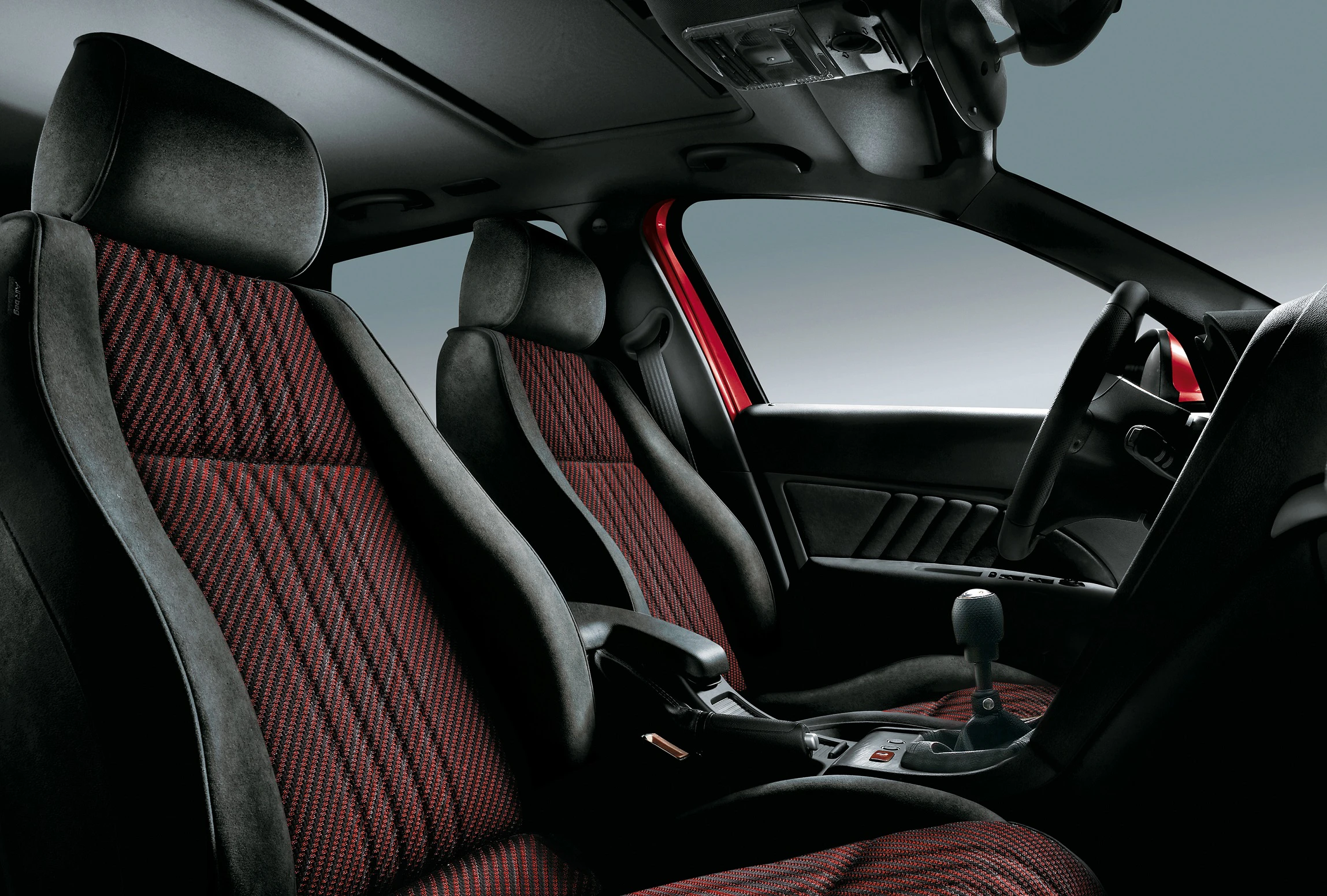Innenraum eines Autos mit roten und schwarzen Sitzbezügen, sportliches Design. Schalthebel in der Mittelkonsole, Dachhimmel schwarz.