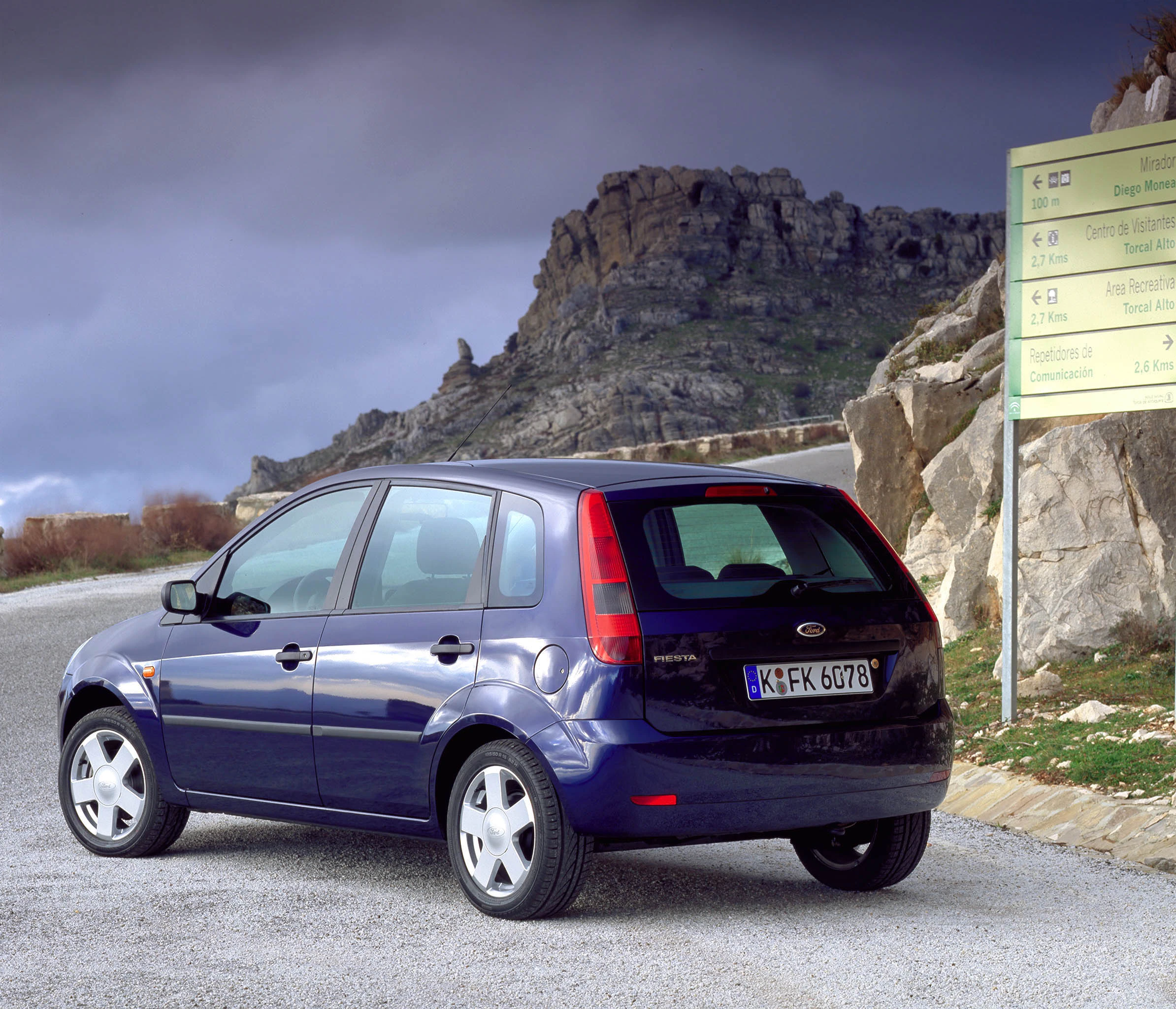 Ein blauer Ford Fiesta parkt auf einer Landstraße bei bewölktem Himmel, im Hintergrund sind Felsen sichtbar. Ein Wegweiser steht rechts im Bild.