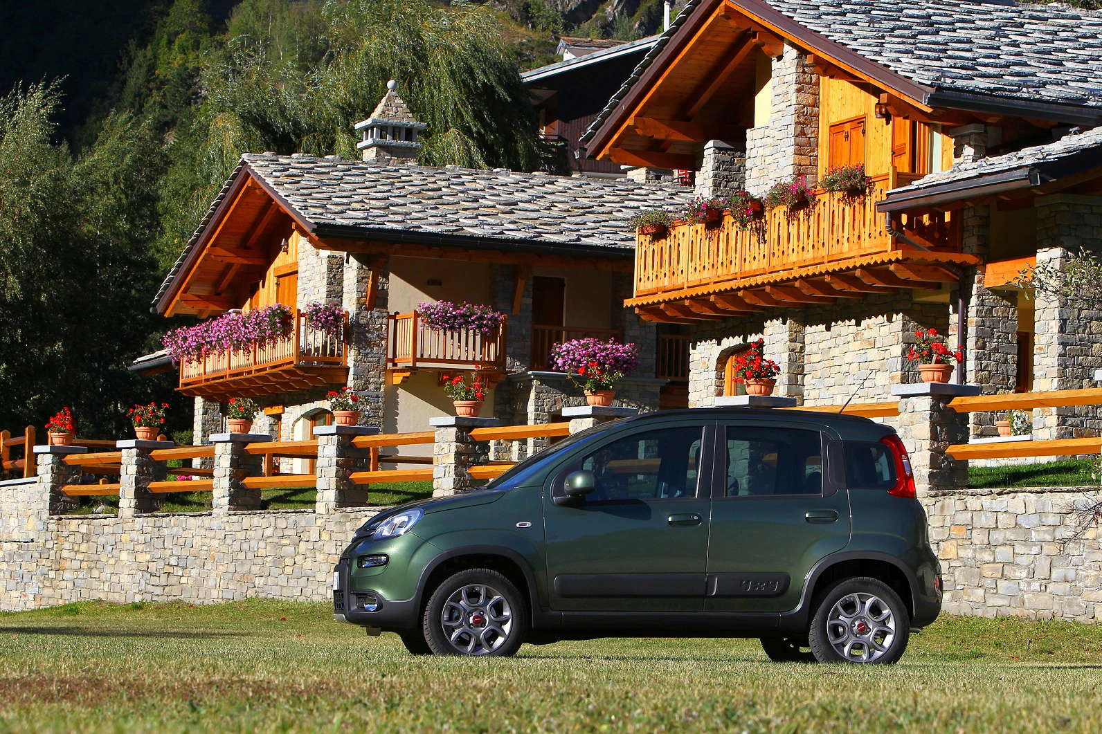 Ein grauer Kleinwagen parkt vor traditionellen Berghütten aus Stein und Holz mit Blumen auf den Balkonen in einer grünen Landschaft.