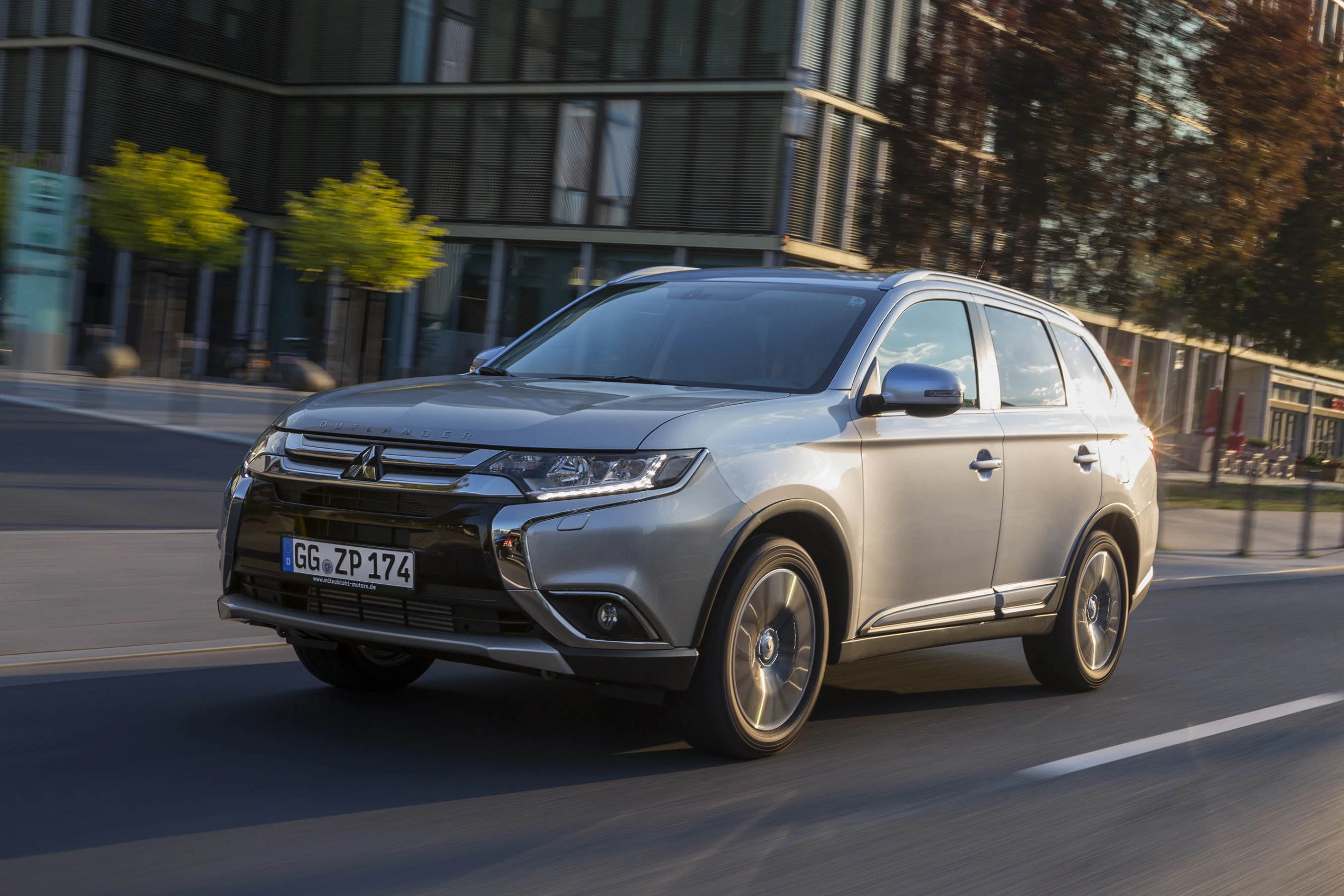 Silberner SUV fährt auf einer Stadtstraße, modernes Design, bei Tageslicht. Im Hintergrund sind Bürogebäude und Bäume zu sehen.