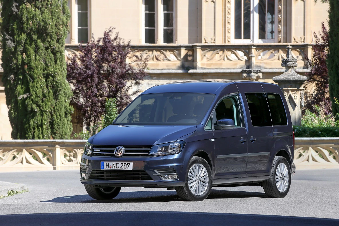 Ein blauer Volkswagen Caddy parkt vor einem historischen Gebäude mit Verzierungen und Bäumen im Hintergrund.