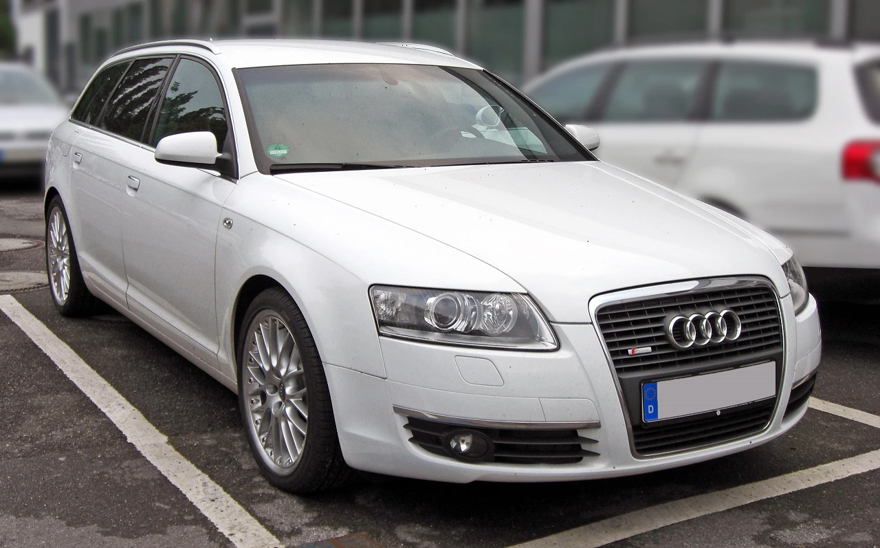 Weißer Audi-Kombi auf einem Parkplatz, technische Details und Emblem sichtbar, umgeben von anderen geparkten Autos.