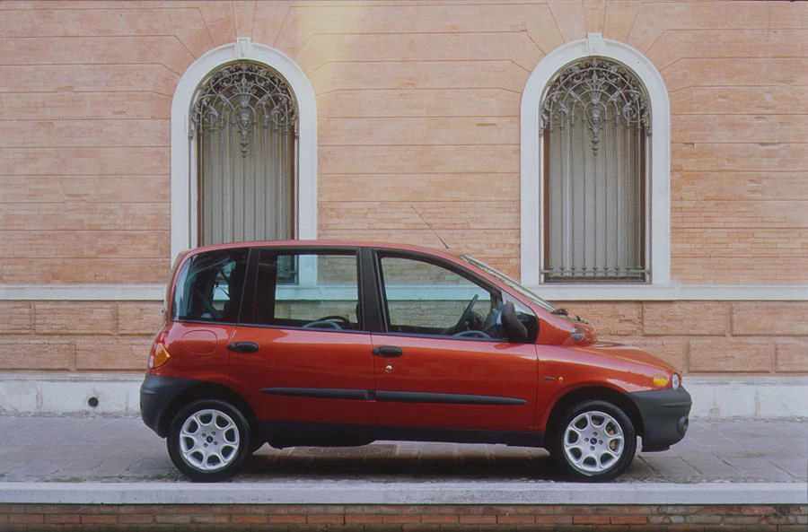 Rotes Auto, geparkt vor einer Backsteinmauer mit zwei verzierten Fenstern. Seitenansicht, klare Linien, kompakt und urban.