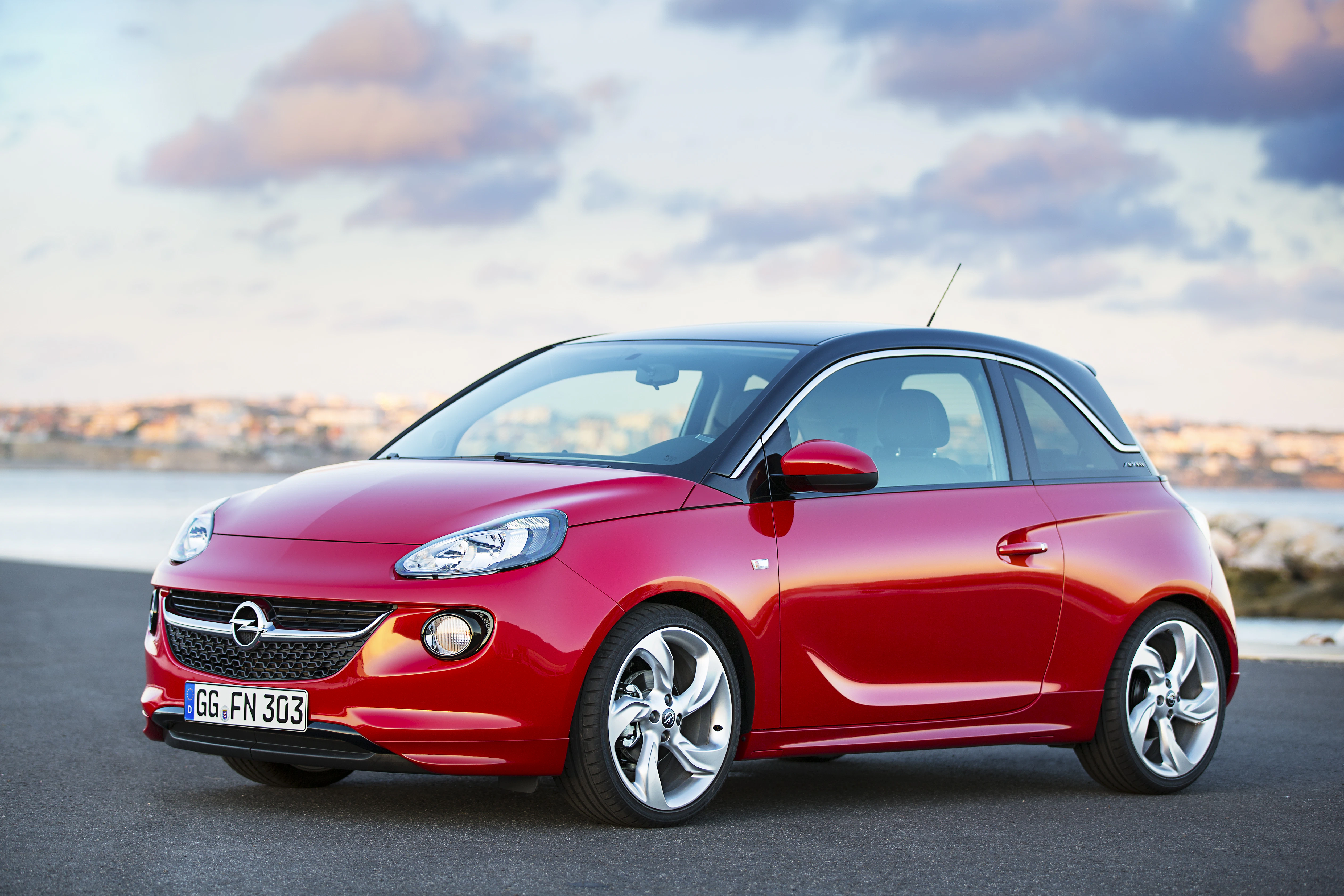Roter Opel Adam auf einer Küstenstraße bei bewölktem Himmel. Kompakte Bauweise, moderne Felgen, Meer im Hintergrund.