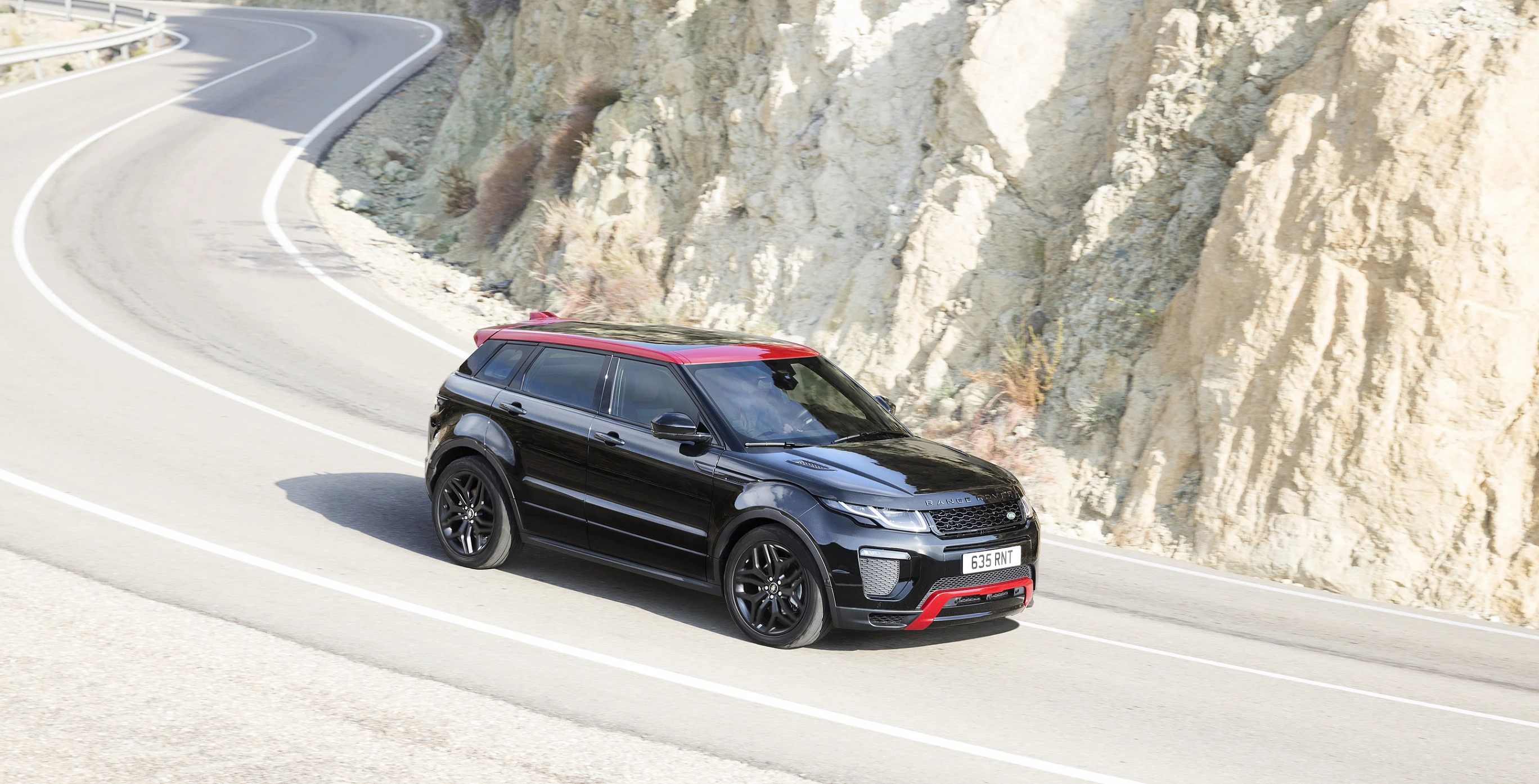 Schwarzer SUV mit rotem Dach fährt auf kurviger Straße entlang einer felsigen Landschaft.