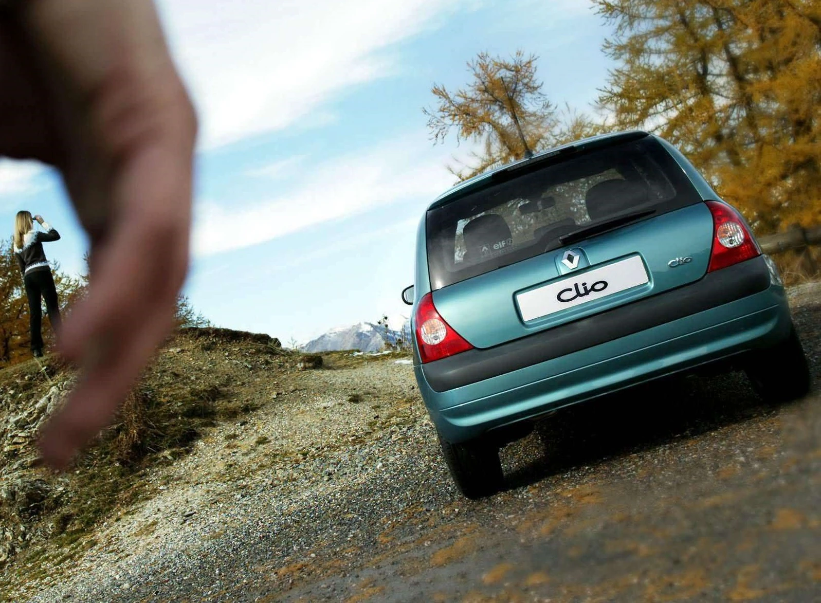 Ein blauer Renault Clio auf einem Schotterweg in einer Berglandschaft, im Hintergrund fotografiert eine Frau die Aussicht.