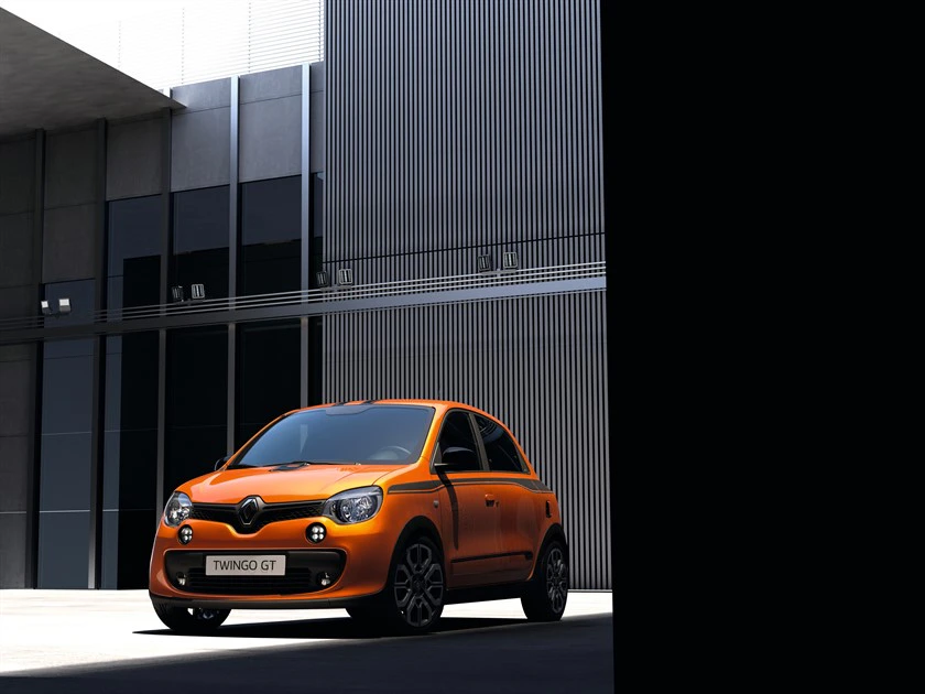 Oranger Renault Twingo GT parkt in einem modernen, schattigen Stadtumfeld mit hohen Gebäuden im Hintergrund.