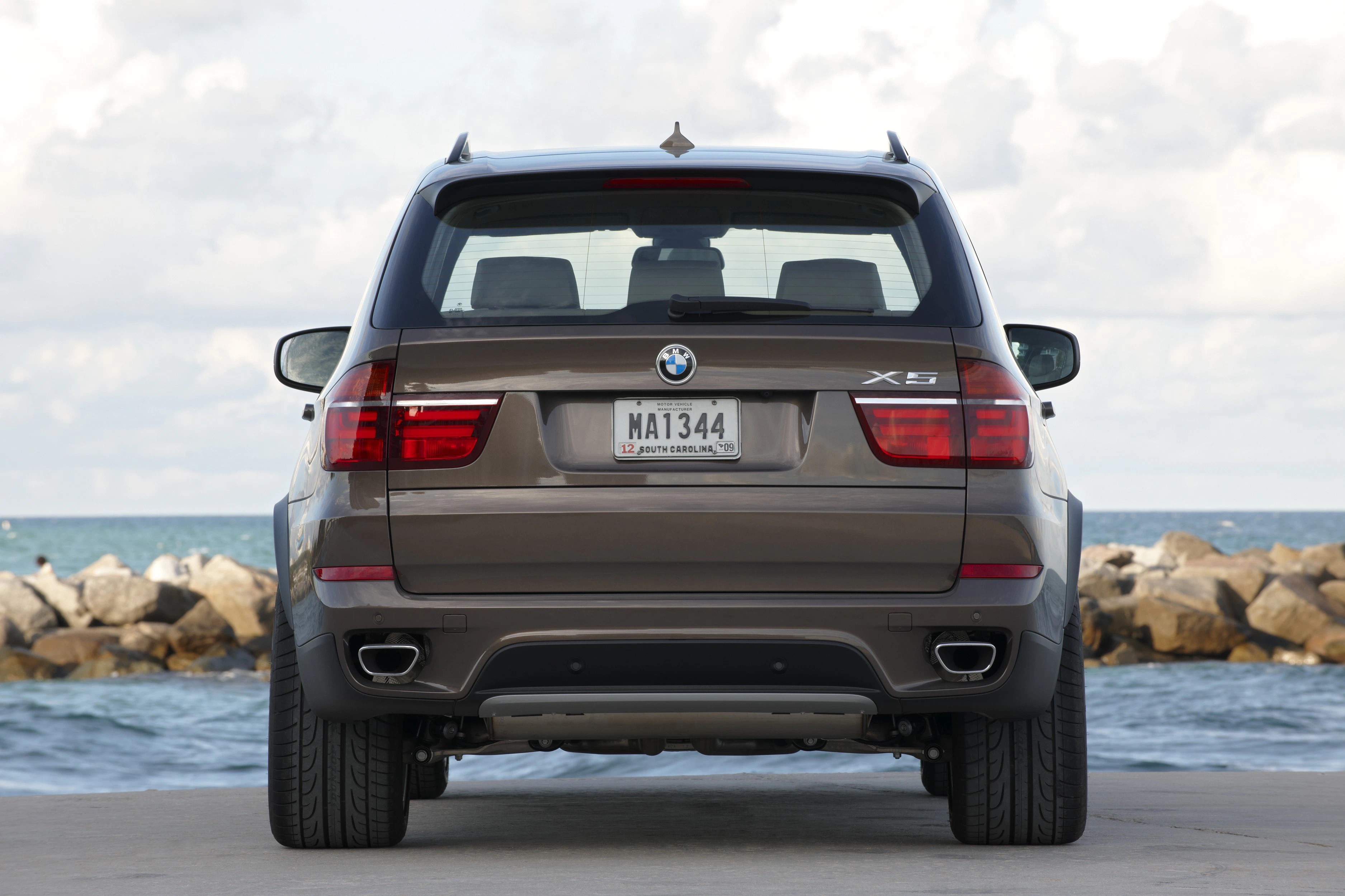 Ein brauner BMW X5 steht am Meer vor Felsen und bewölktem Himmel, von hinten fotografiert.