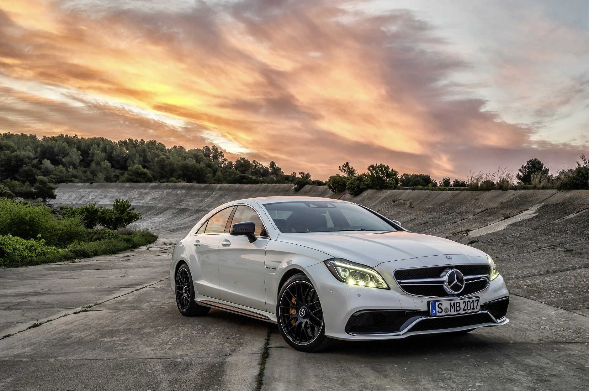 Weiße Limousine von Mercedes-Benz auf einer Betonfläche bei Sonnenuntergang, umgeben von Bäumen und einem farbenfrohen Himmel.