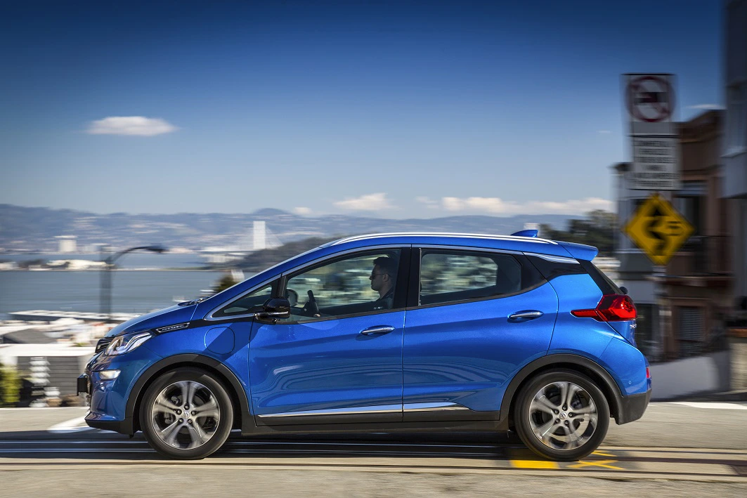 Blaues Elektroauto fährt am Tag eine sanfte Steigung hinauf; Stadt und Wasser im Hintergrund sichtbar.
