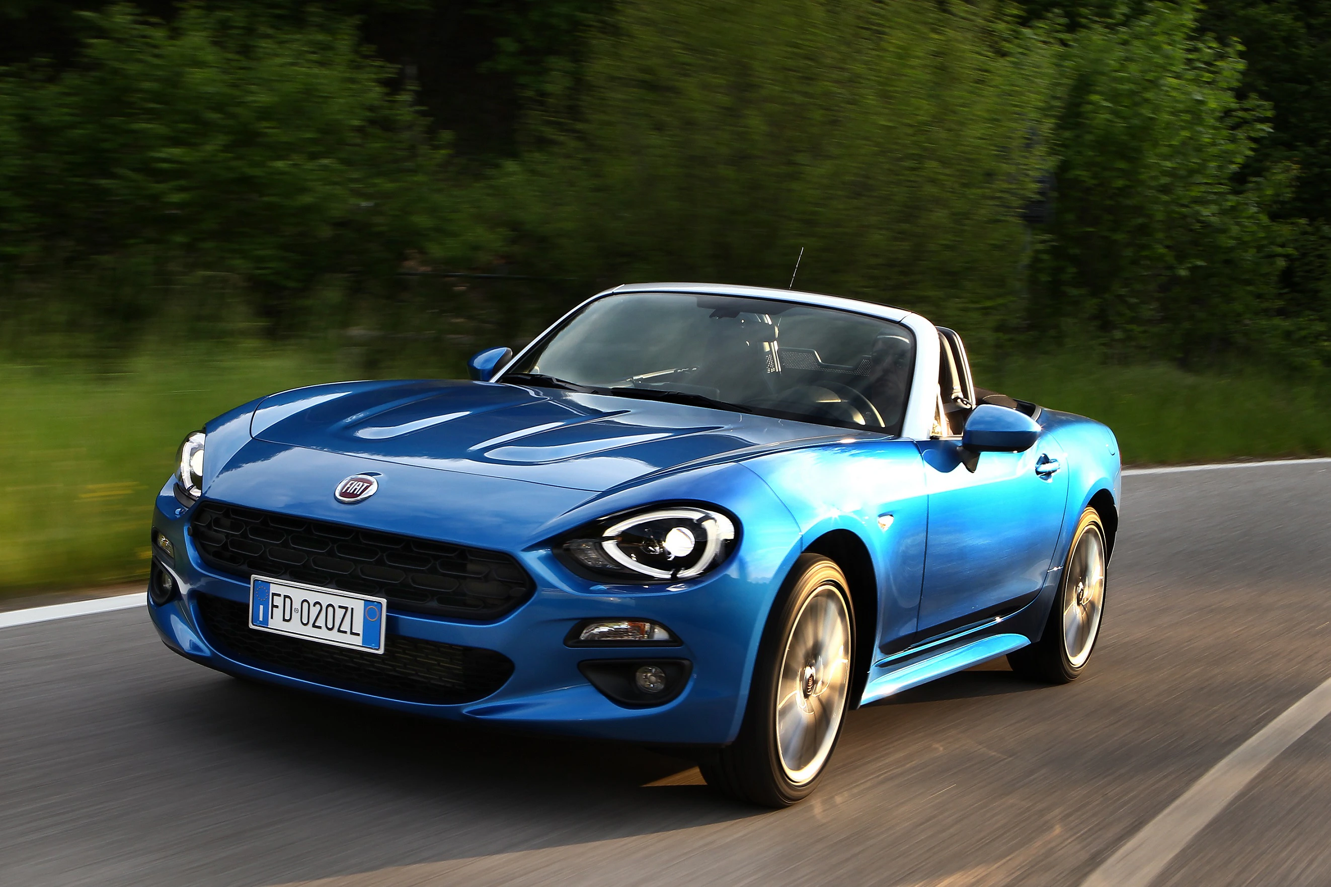 Blauer Sportwagen auf einer Landstraße bei Tageslicht, umgeben von grüner Natur. Das Auto hat ein Cabrio-Dach und fährt mit hoher Geschwindigkeit.