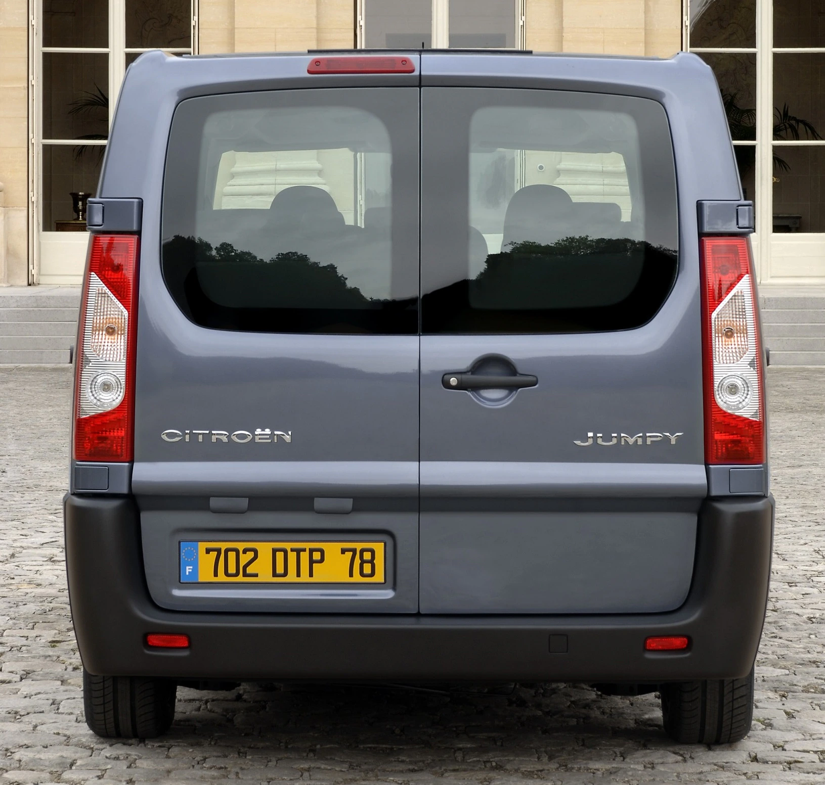 Heckansicht eines grauen Citroën Jumpy, mit französischem Kennzeichen, geparkt auf Pflastersteinen vor einem Gebäude mit Treppe und Fenstern.