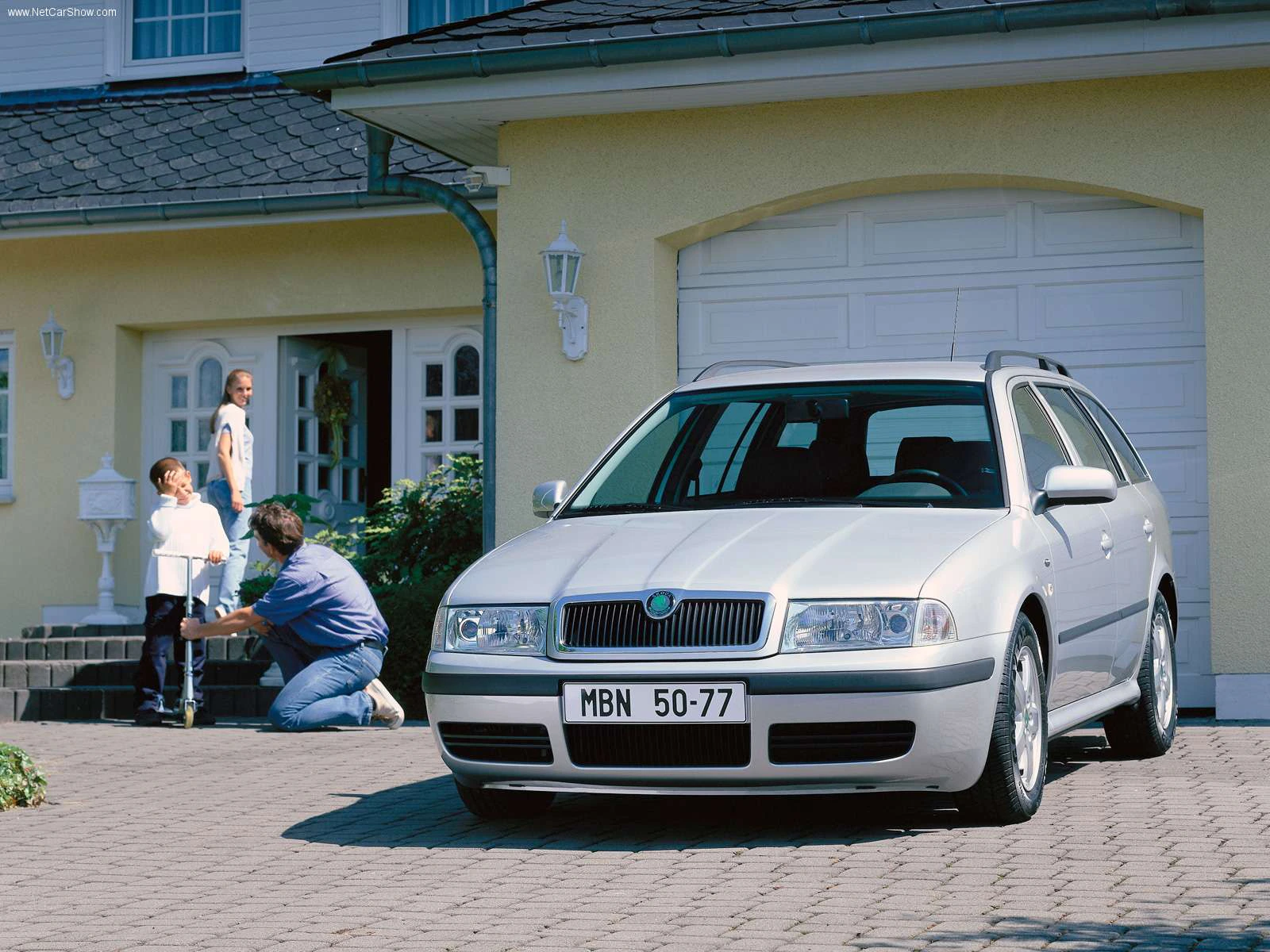 Silberner Kombi auf einer Einfahrt vor einem gelben Haus, im Hintergrund eine Familie mit zwei Kindern.