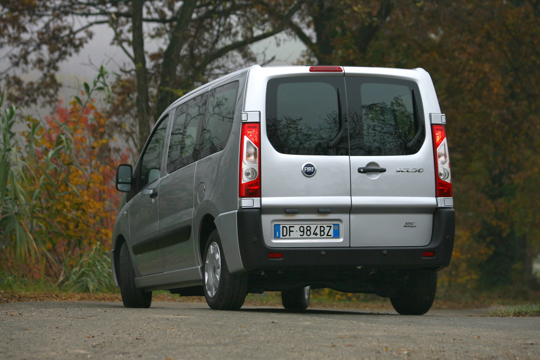 Silberner Fiat-Kleinbus, geparkt auf einer Straße, umgeben von herbstlicher Landschaft und Bäumen im Hintergrund.
