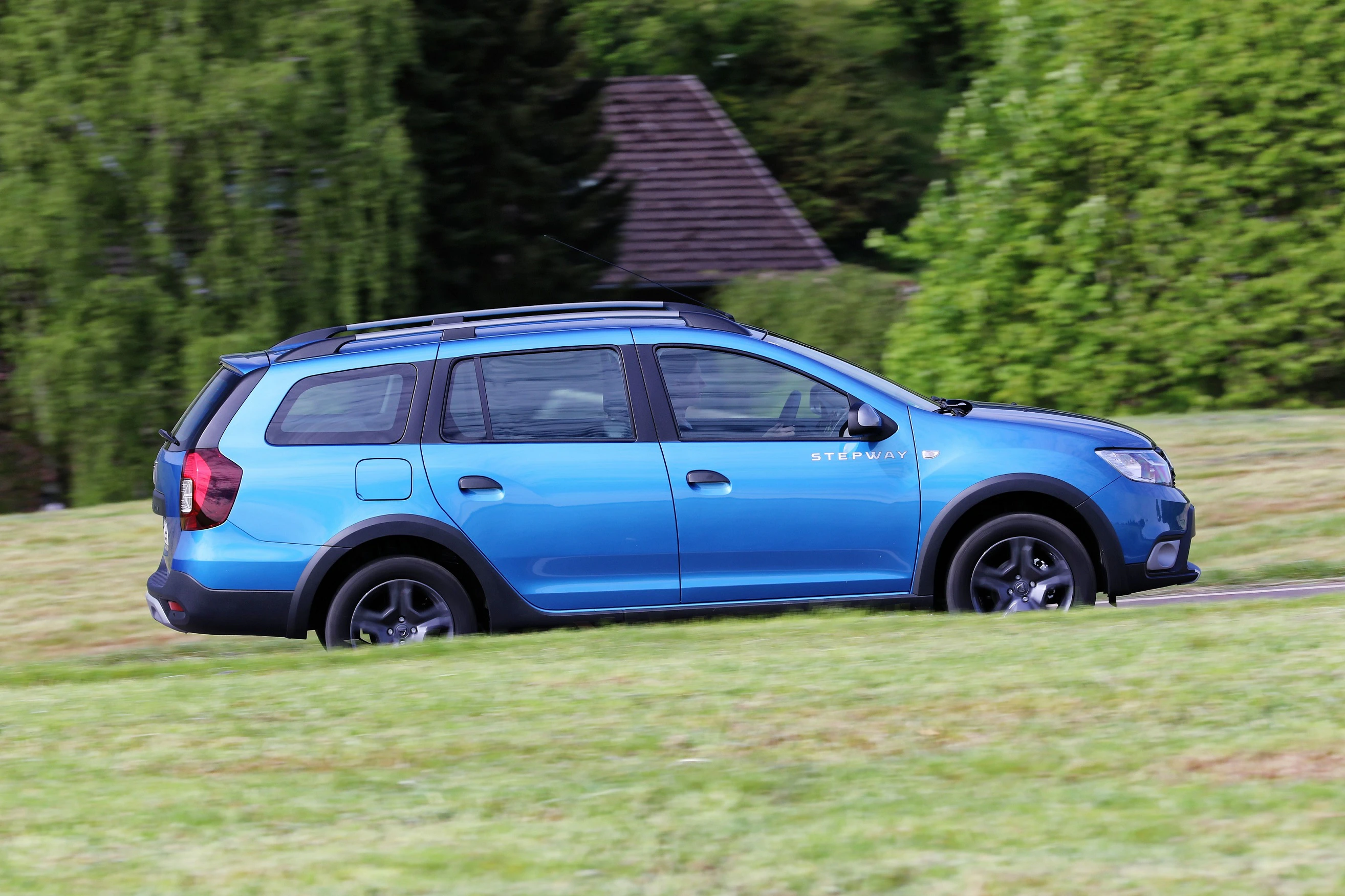 Blauer Kombi fährt auf einer grünen Wiese vor einem Hintergrund mit Bäumen und einem Hausdach.