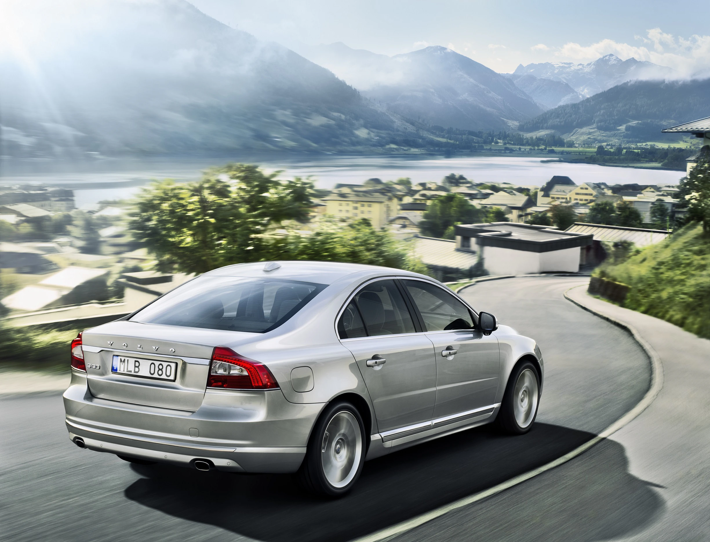Silberner Volvo S80 fährt auf einer kurvigen Straße mit Blick auf eine malerische Landschaft aus Bergen und einem See bei sonnigem Wetter.